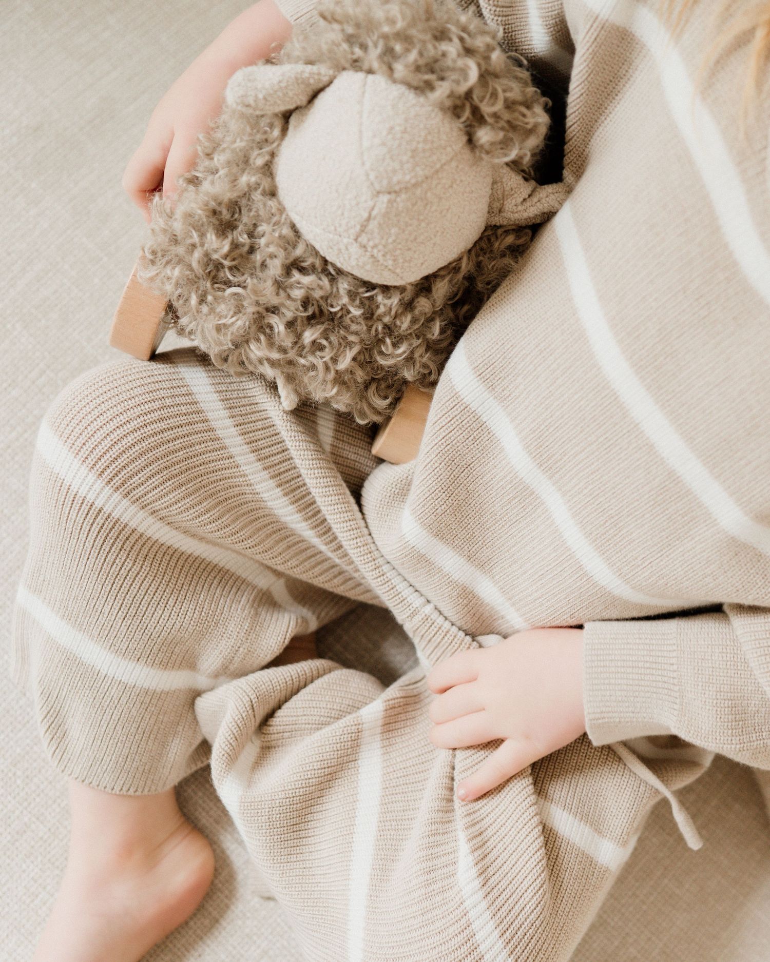 Child in ribbed beige outfit cuddling a plush lamb toy under a striped knit blanket.
