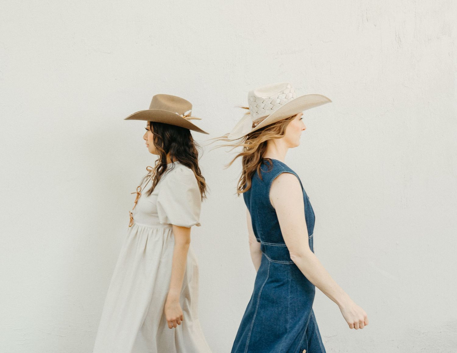 Two women in cowboy hats and boho dresses pose back-to-back against a white wall.