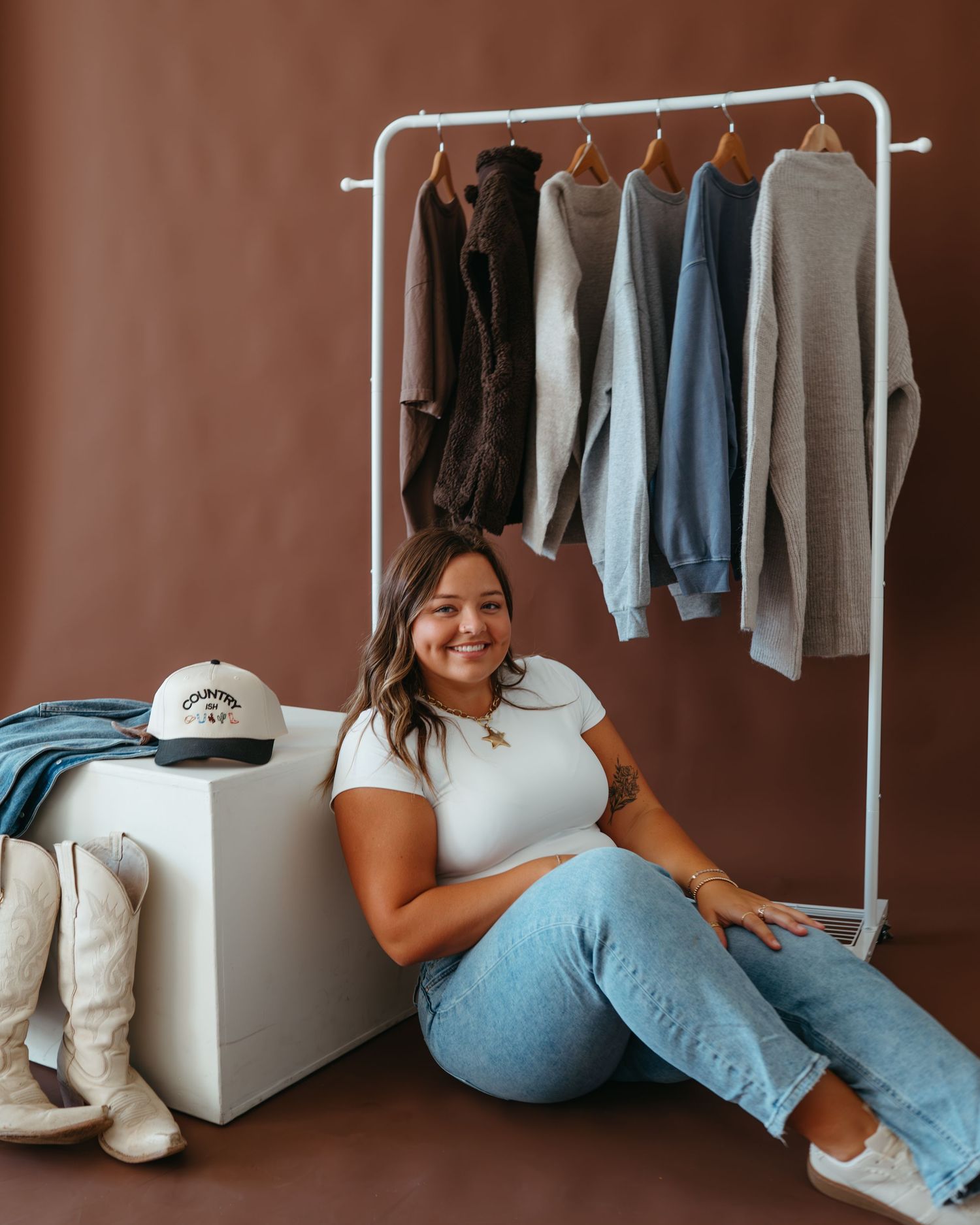 Smiling woman in white tee and jeans sitting beside a clothing rack with neutral-toned sweatshirts on display.