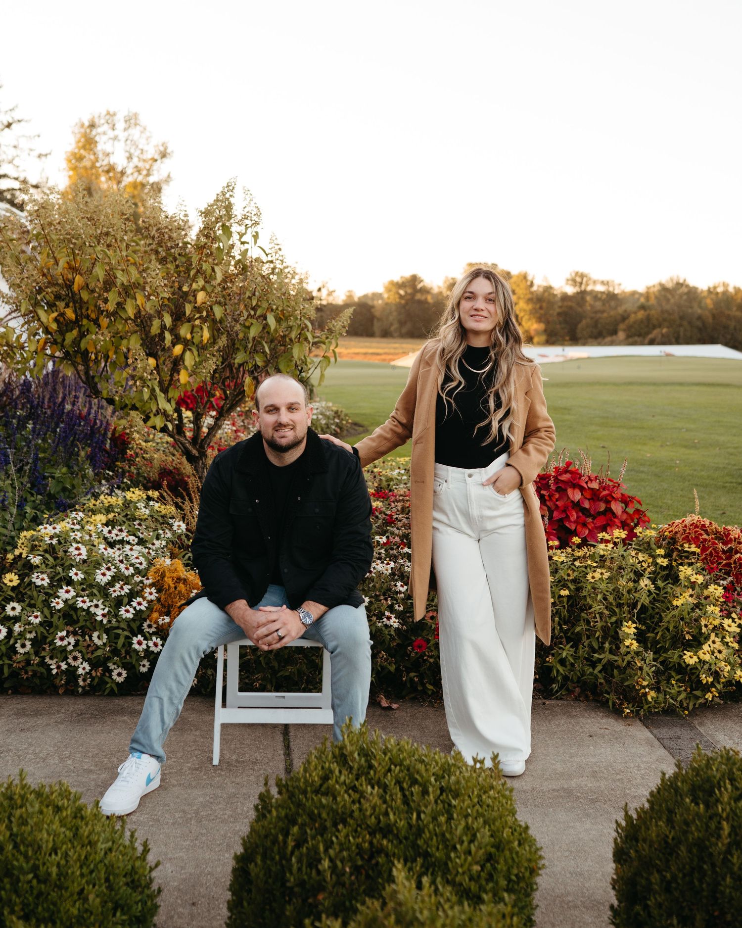 Smiling couple posing among colorful fall flowers at a scenic golf course garden during a warm sunset evening.