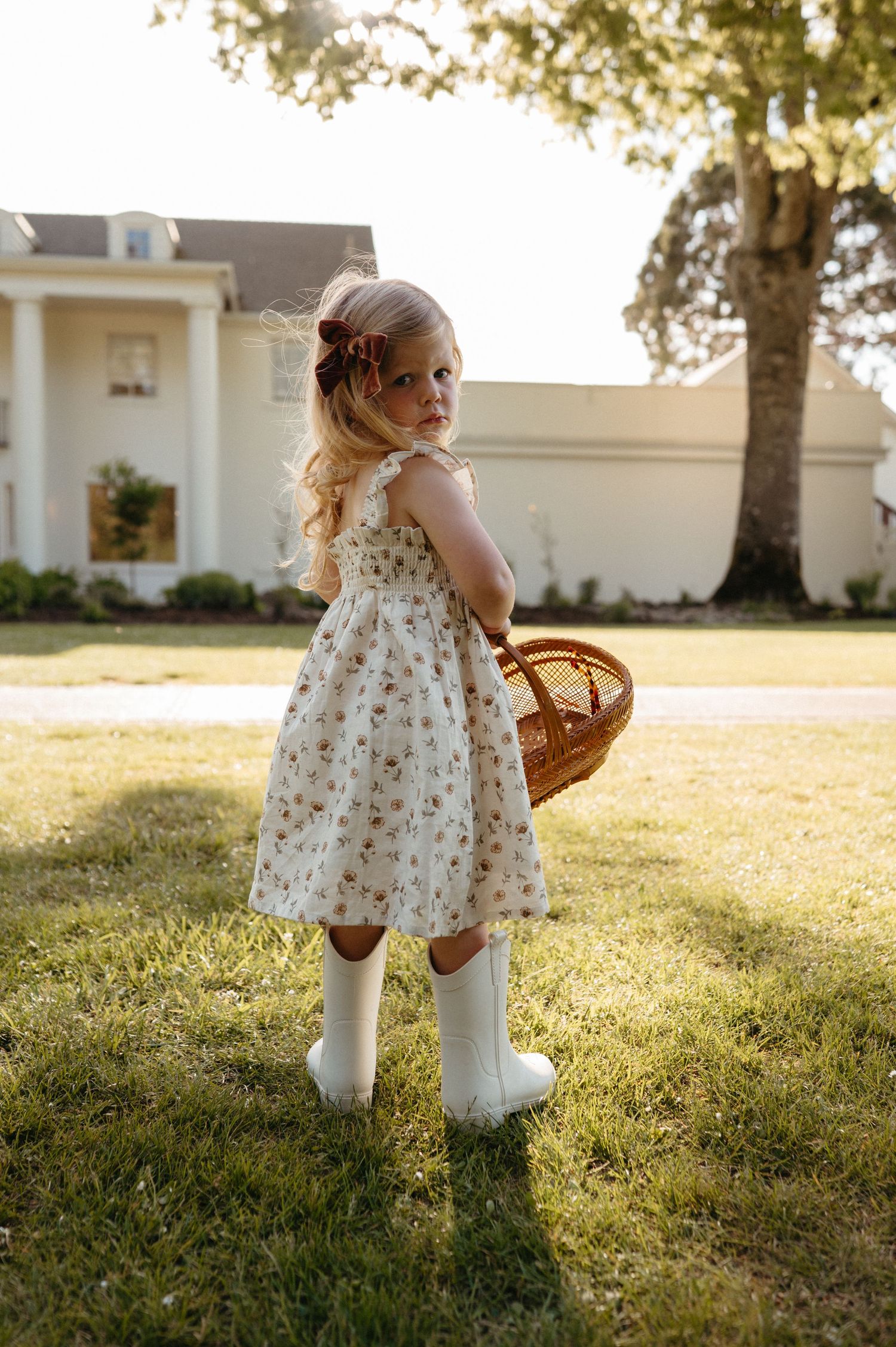 Young girl in floral dress and white boots holding a wicker basket outdoors near a white house.