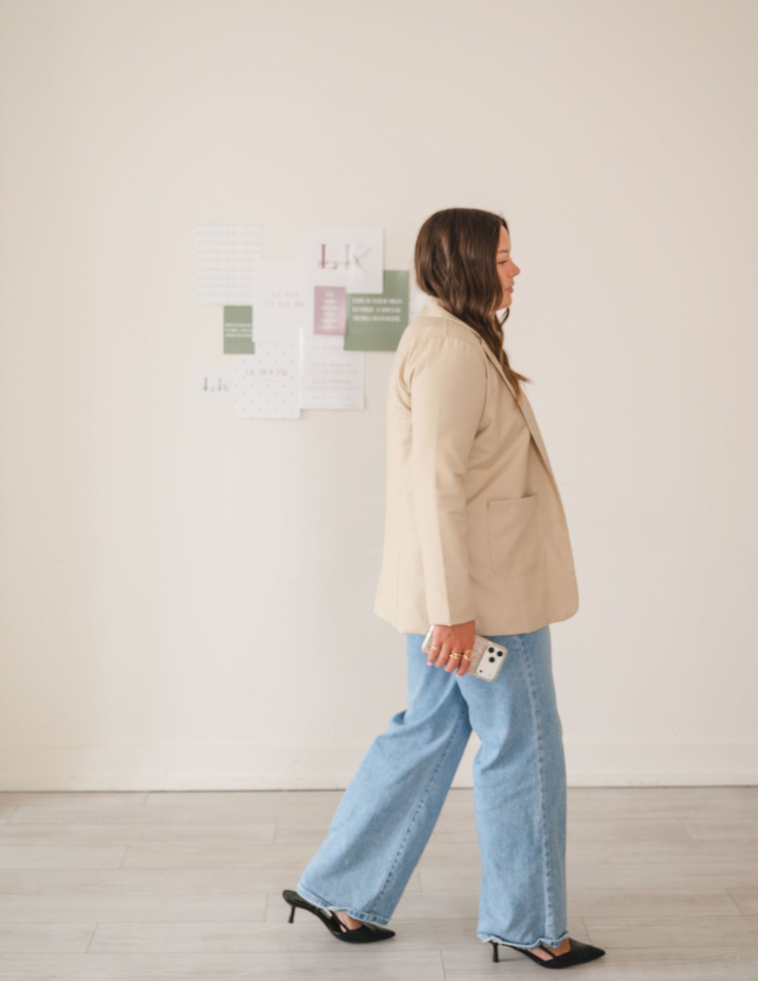 Woman in beige blazer and wide-leg jeans walking past a mood board on a white wall.