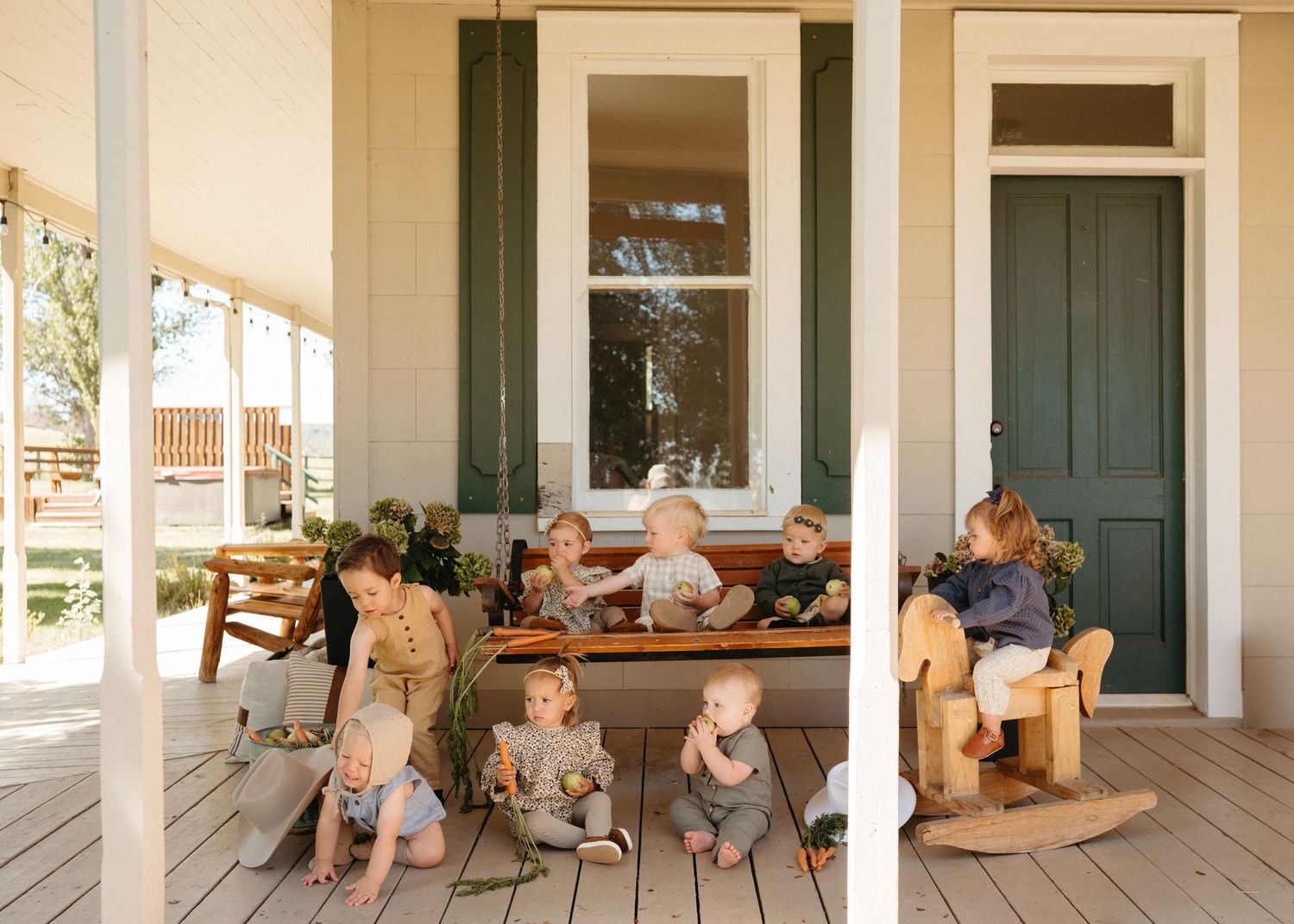 Several toddlers and young children gathered on a charming beige farmhouse porch with green shutters.