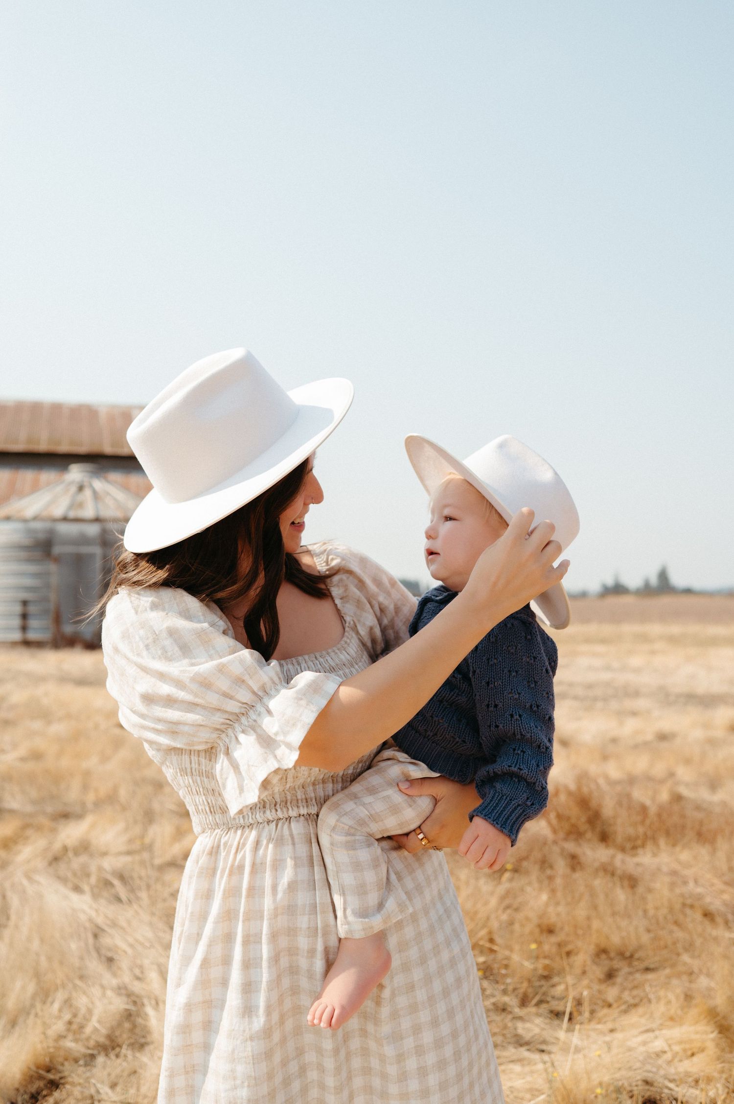 Mother in white hat holds baby with matching hat in a golden wheat field near a barn.