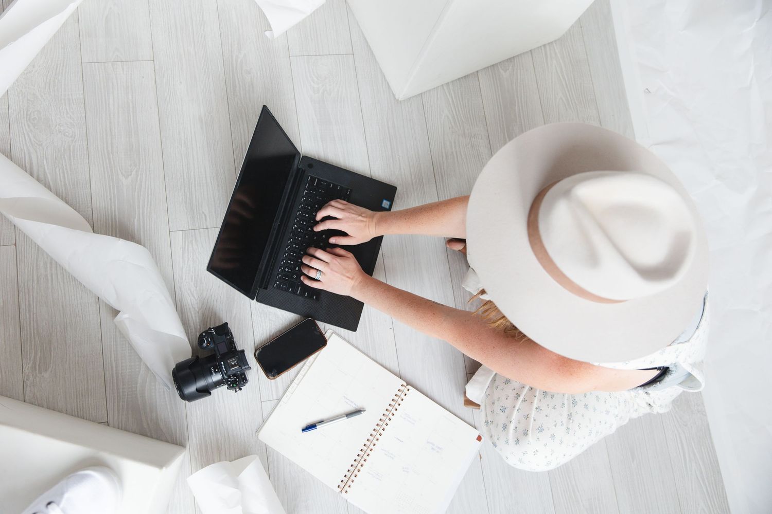 Overhead view of a woman in a white hat working on a laptop on a light wood floor with a camera and notebook nearby.