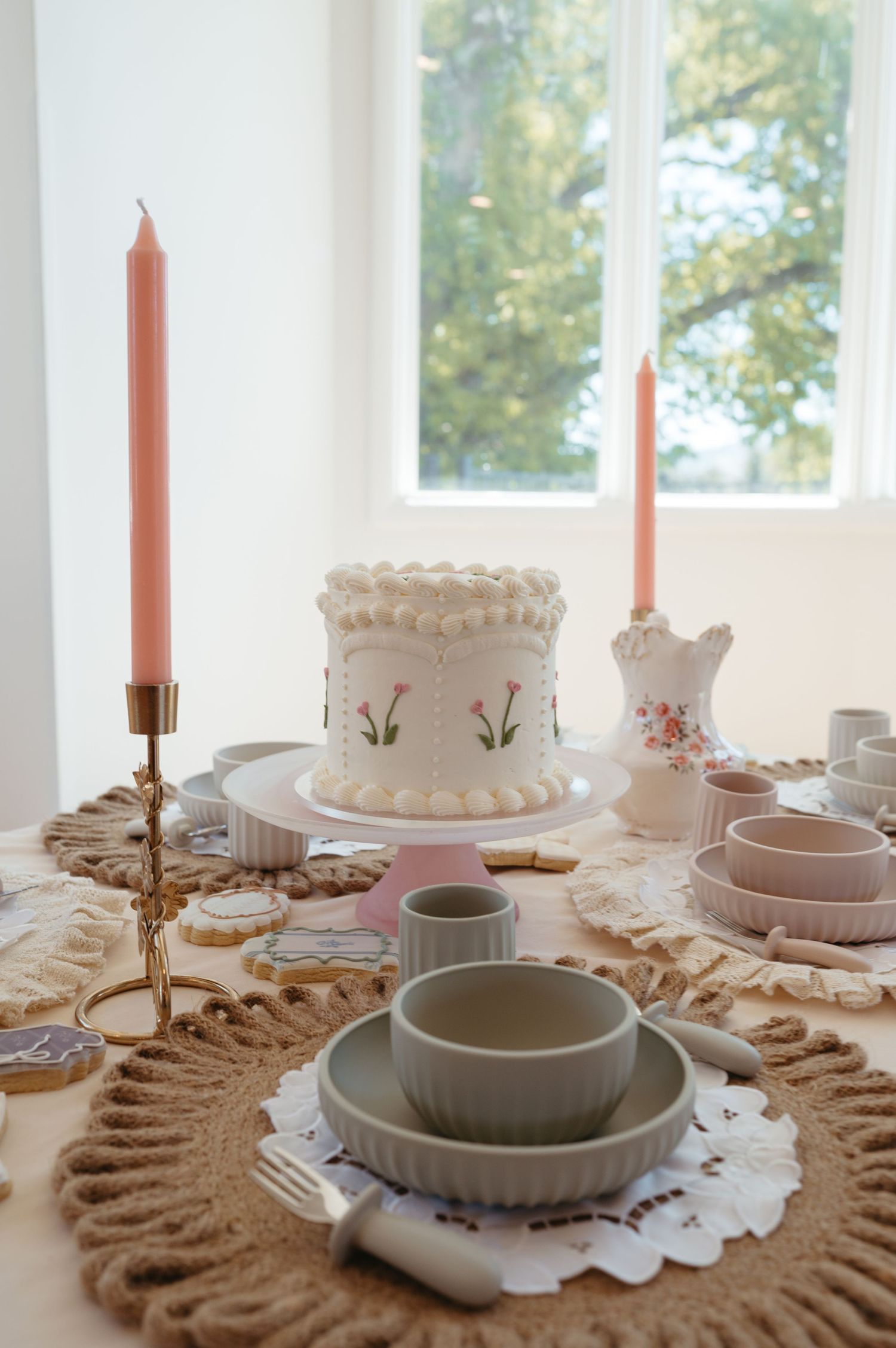 Elegant birthday table with a floral white cake, pink taper candles, and ceramic dishes.
