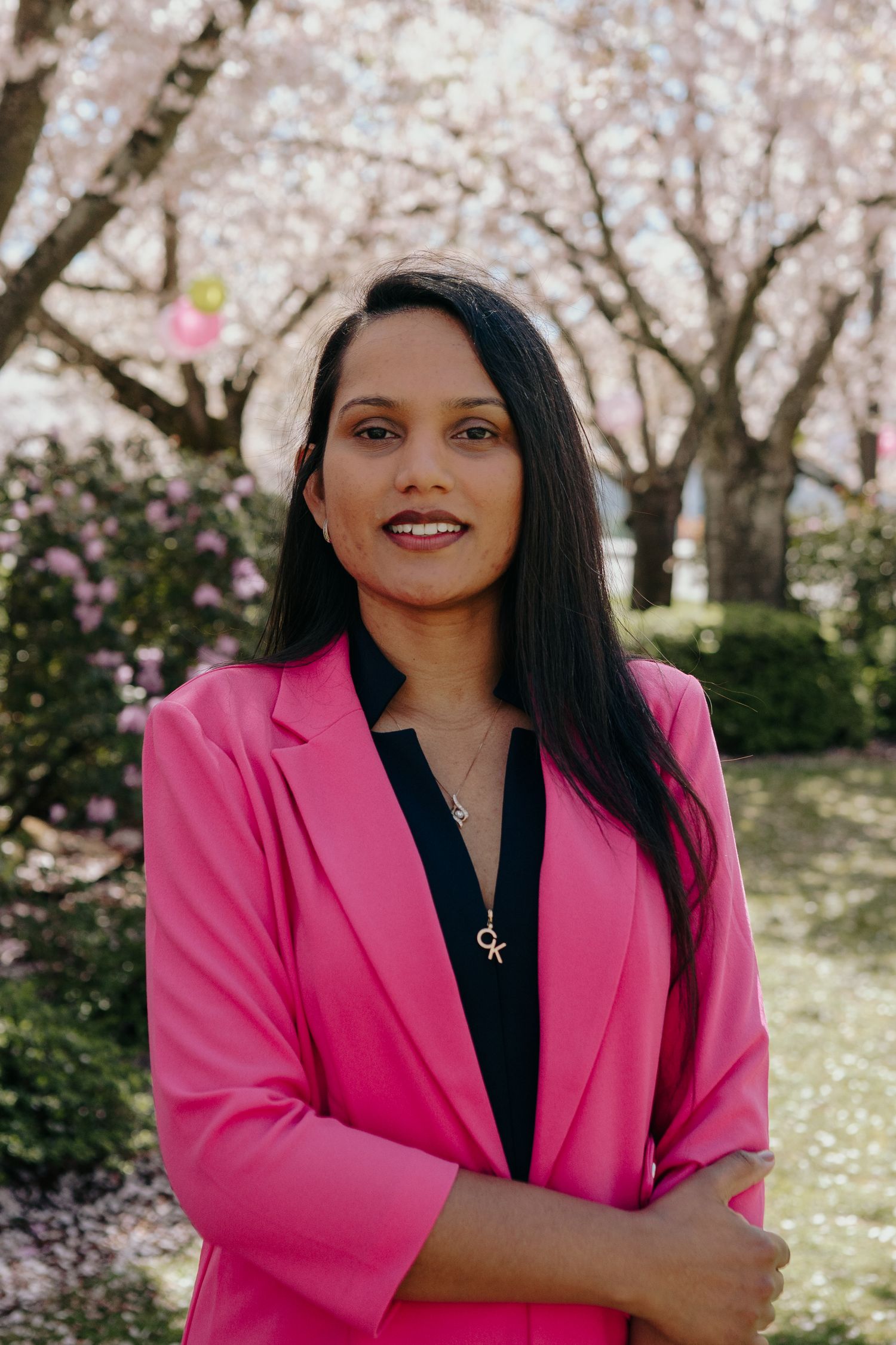 Smiling woman in pink blazer posing outdoors among blooming cherry blossom trees in spring.