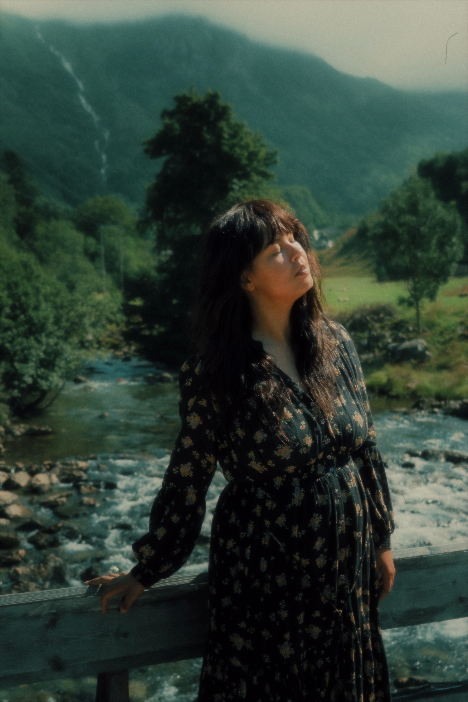 A silhouette stands near a flowing river with mountains and trees in the background.