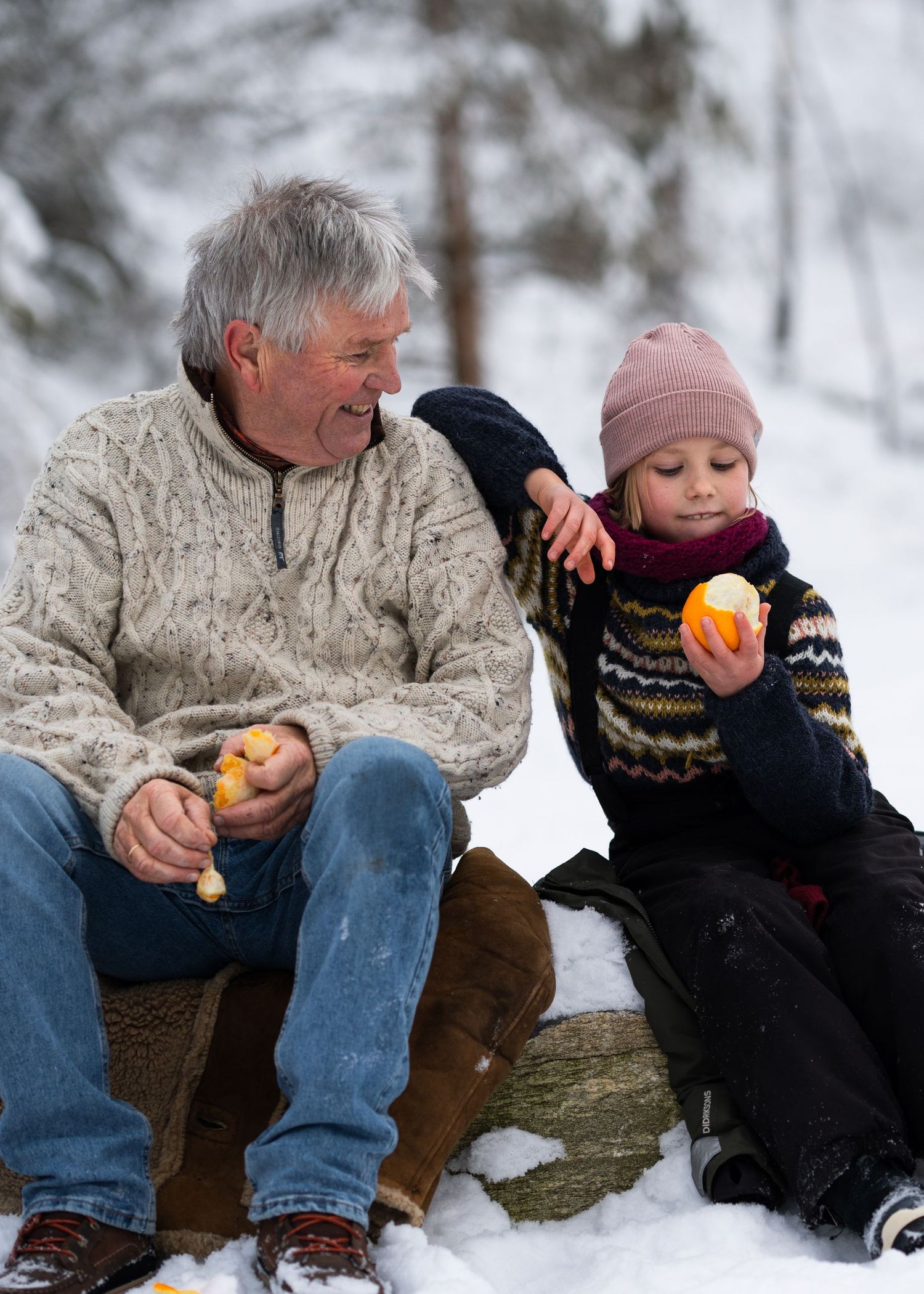 Two people share oranges while sitting in snowy winter landscape.
