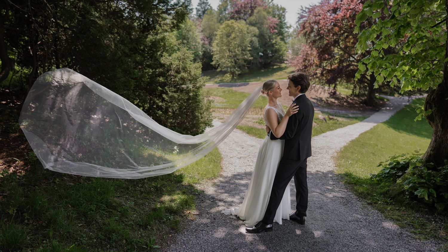 A romantic wedding couple walks down a tree-lined path in a park setting.