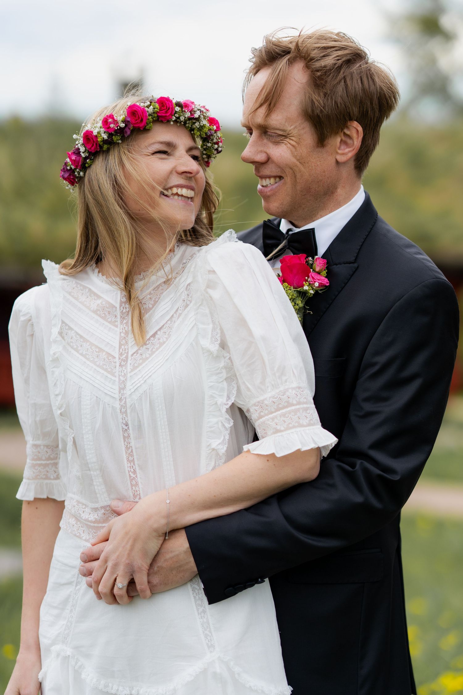 Couple in wedding attire embraces in a meadow with red flower crown details.