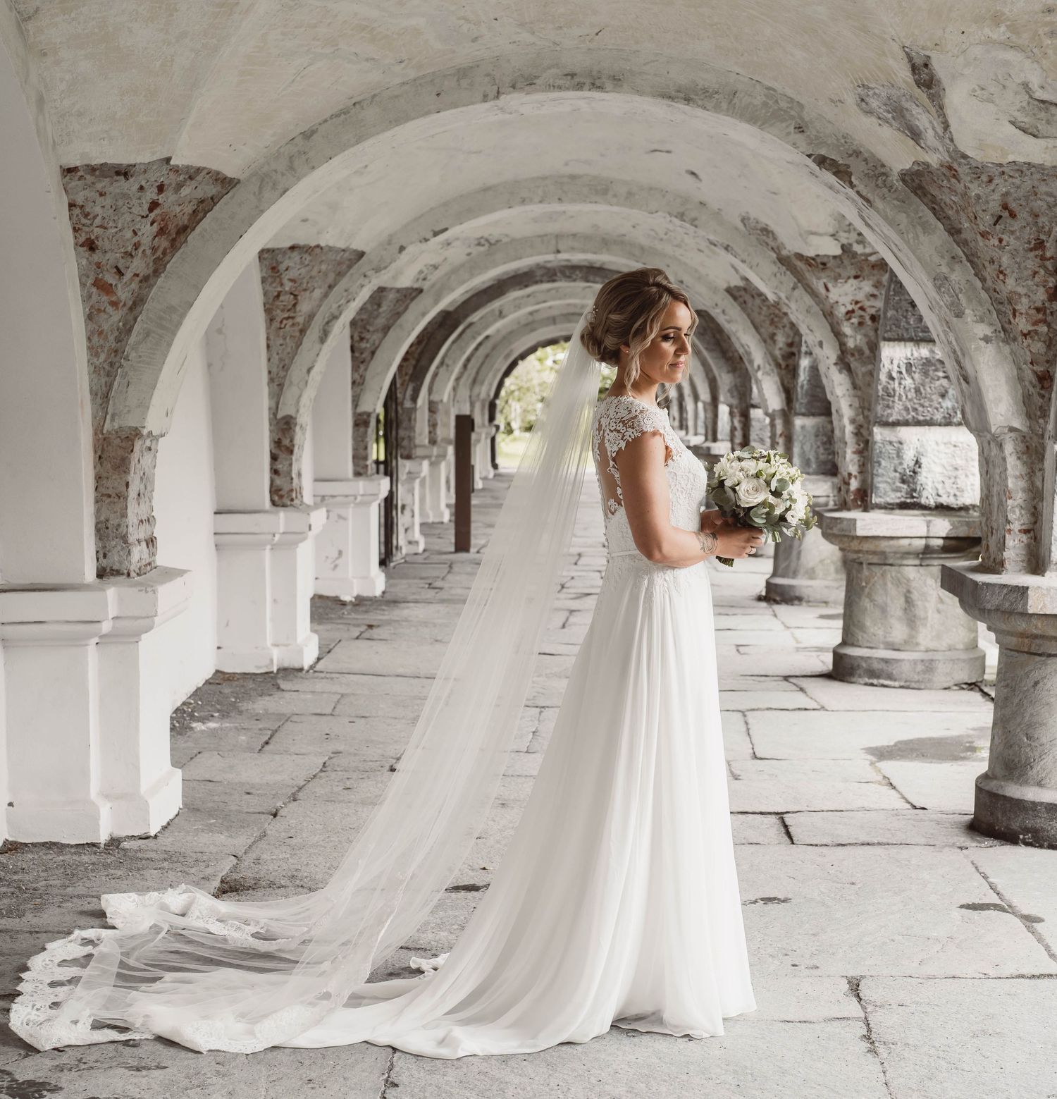 Bride in flowing white dress poses in historic stone archway corridor.