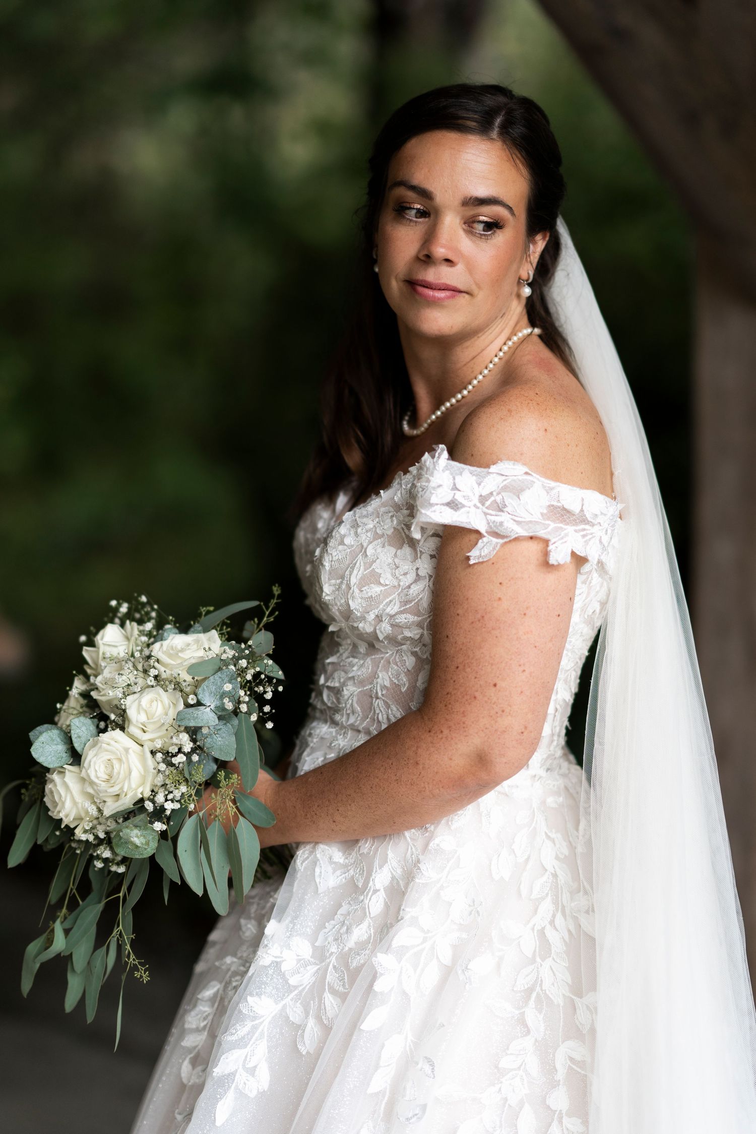 Brud i off-shoulder kjole poserer med en hvit rose- og eukalyptusbukett.