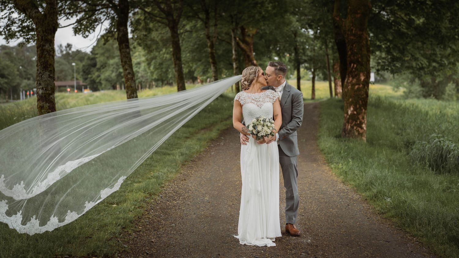 Wedding couple poses on a scenic wooded path with flowing veil in natural lighting.