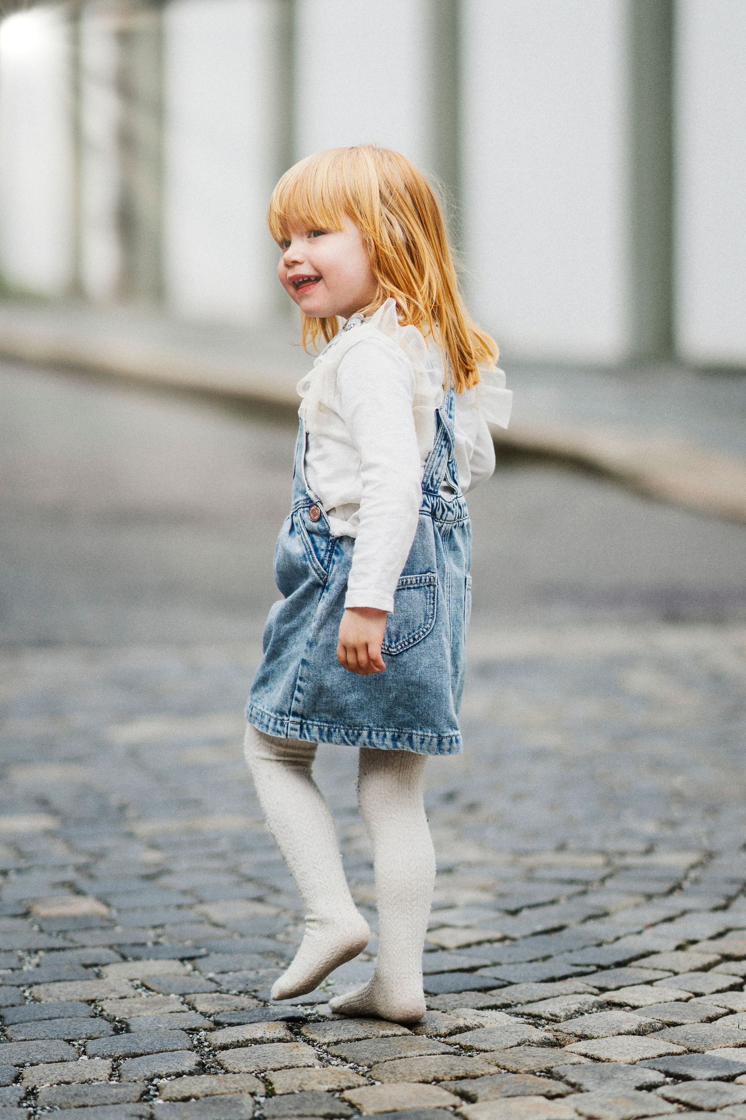 Small child in white tights and denim dress walking on cobblestone street.