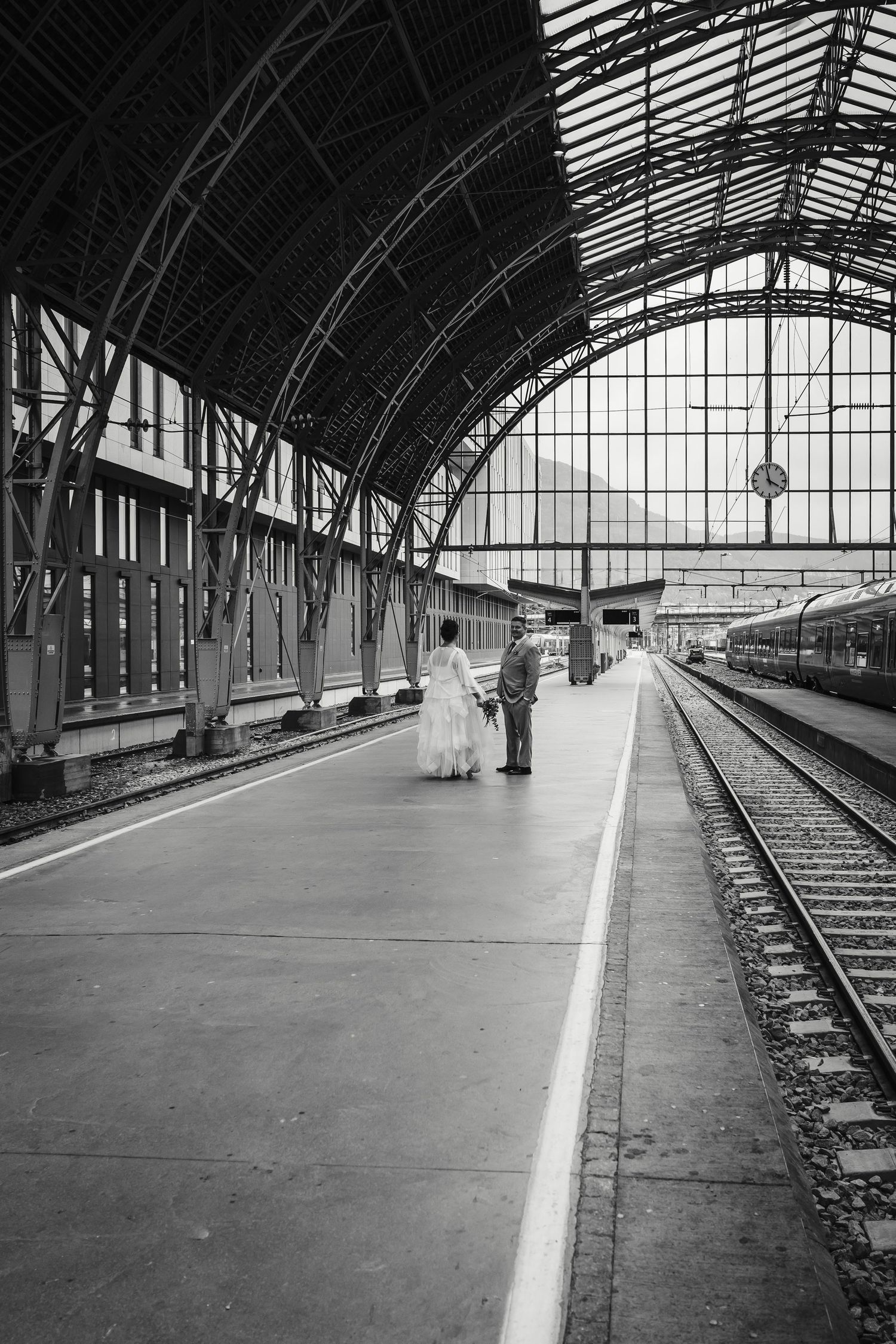 A vintage train station platform with arched glass ceiling and waiting passengers under dramatic architecture.