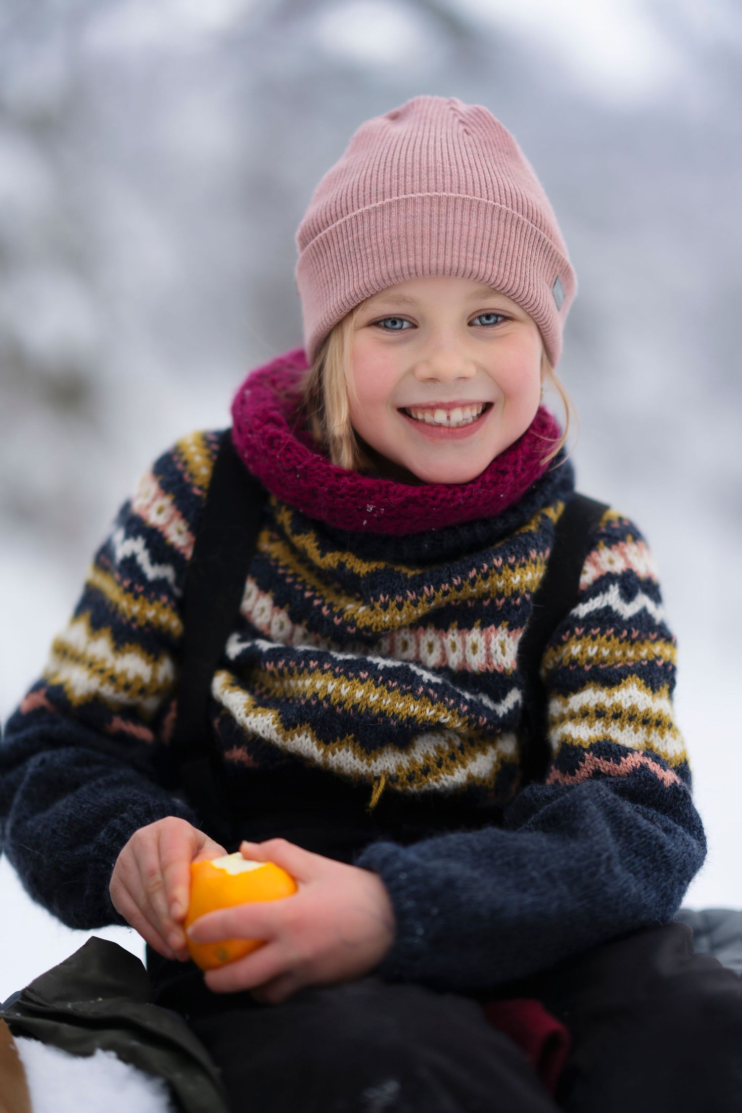 Person in knit sweater and pink hat holds orange fruit in snowy setting.