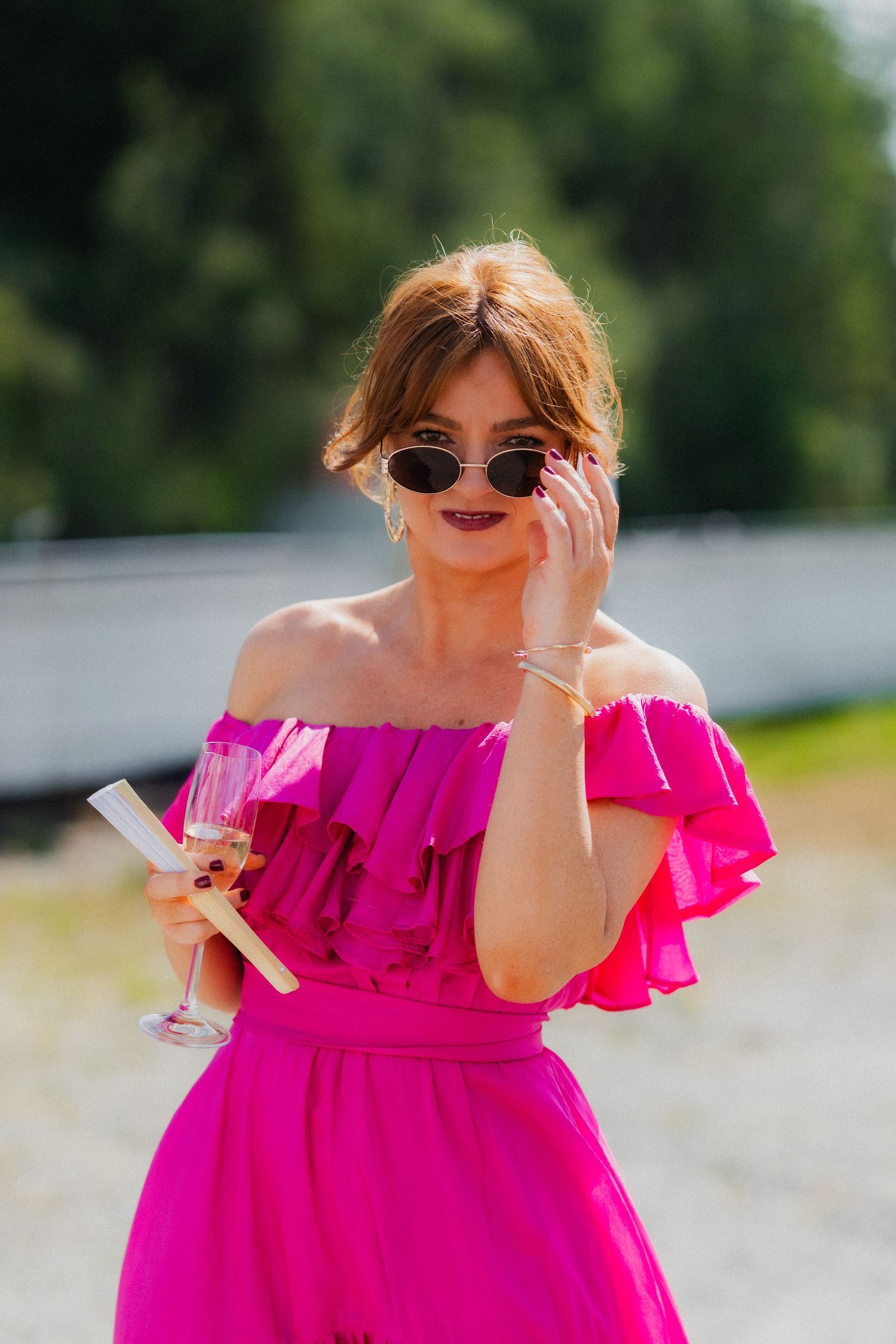 Person in bright pink off-shoulder dress holding drink outdoors wearing sunglasses.