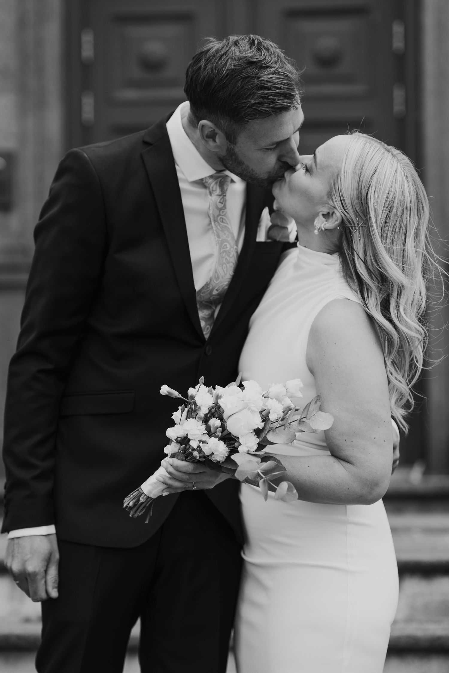 A romantic black and white wedding photo of a couple sharing a kiss while holding a bouquet.