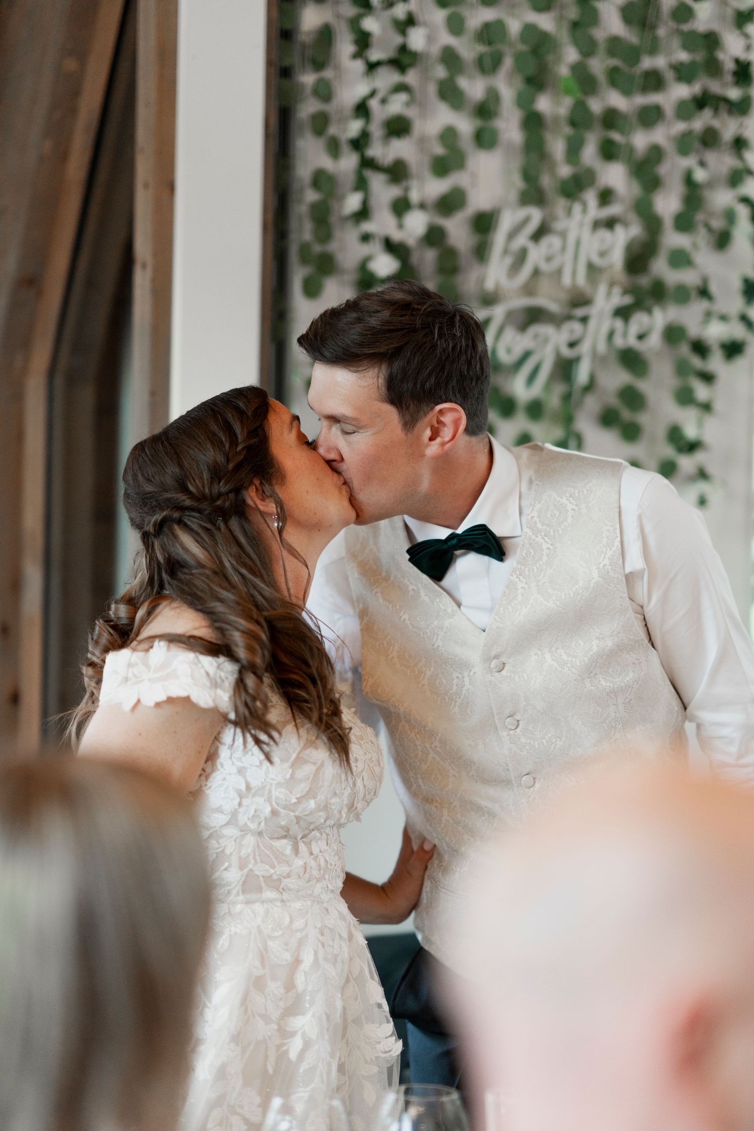 Newlyweds share a romantic kiss during their wedding ceremony with greenery in the background.