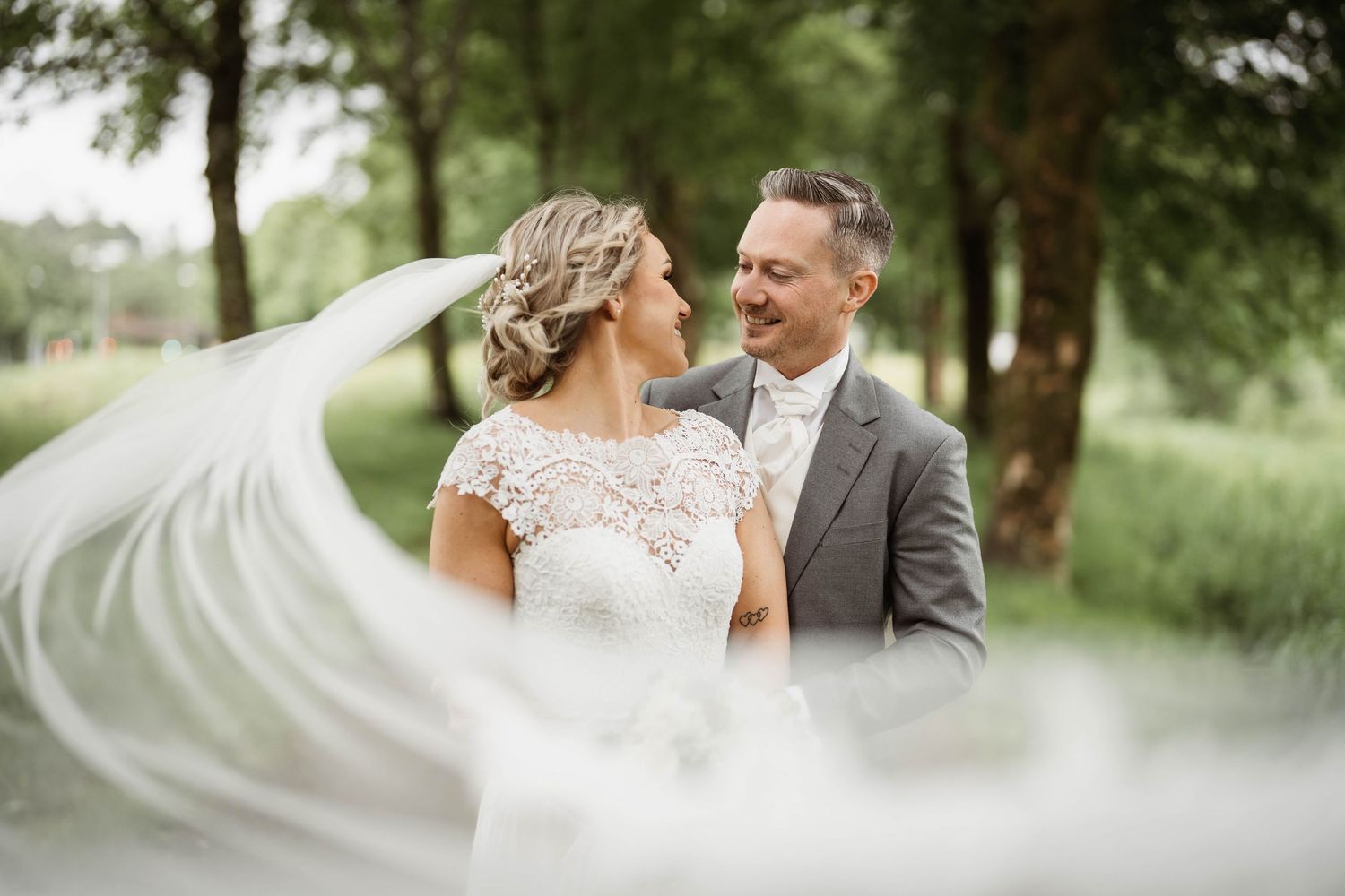 Wedding couple embraces outdoors with bride's veil flowing dramatically in the wind.