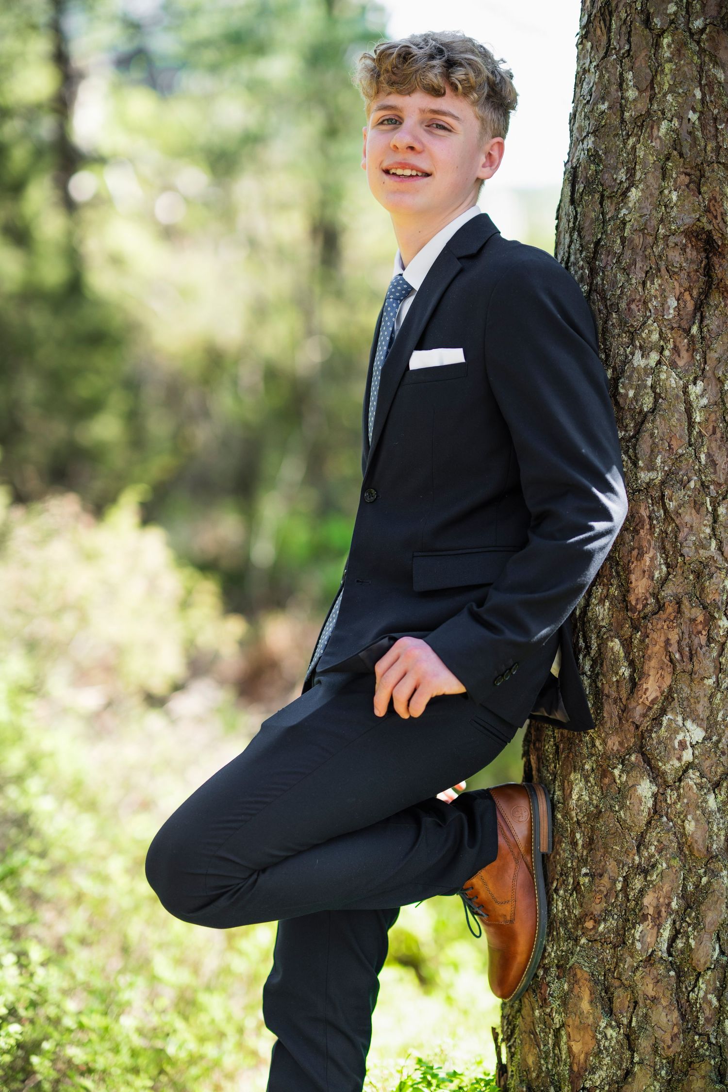 Outdoor portrait poses against a tree trunk wearing a formal black suit in natural lighting.