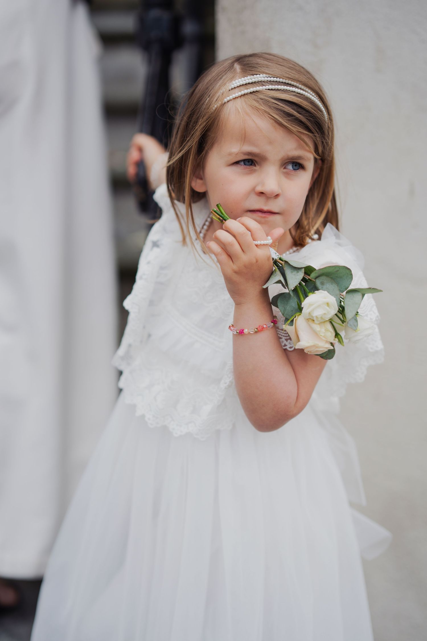 A flower girl in a white lace dress holds a delicate rose bouquet at a formal event.