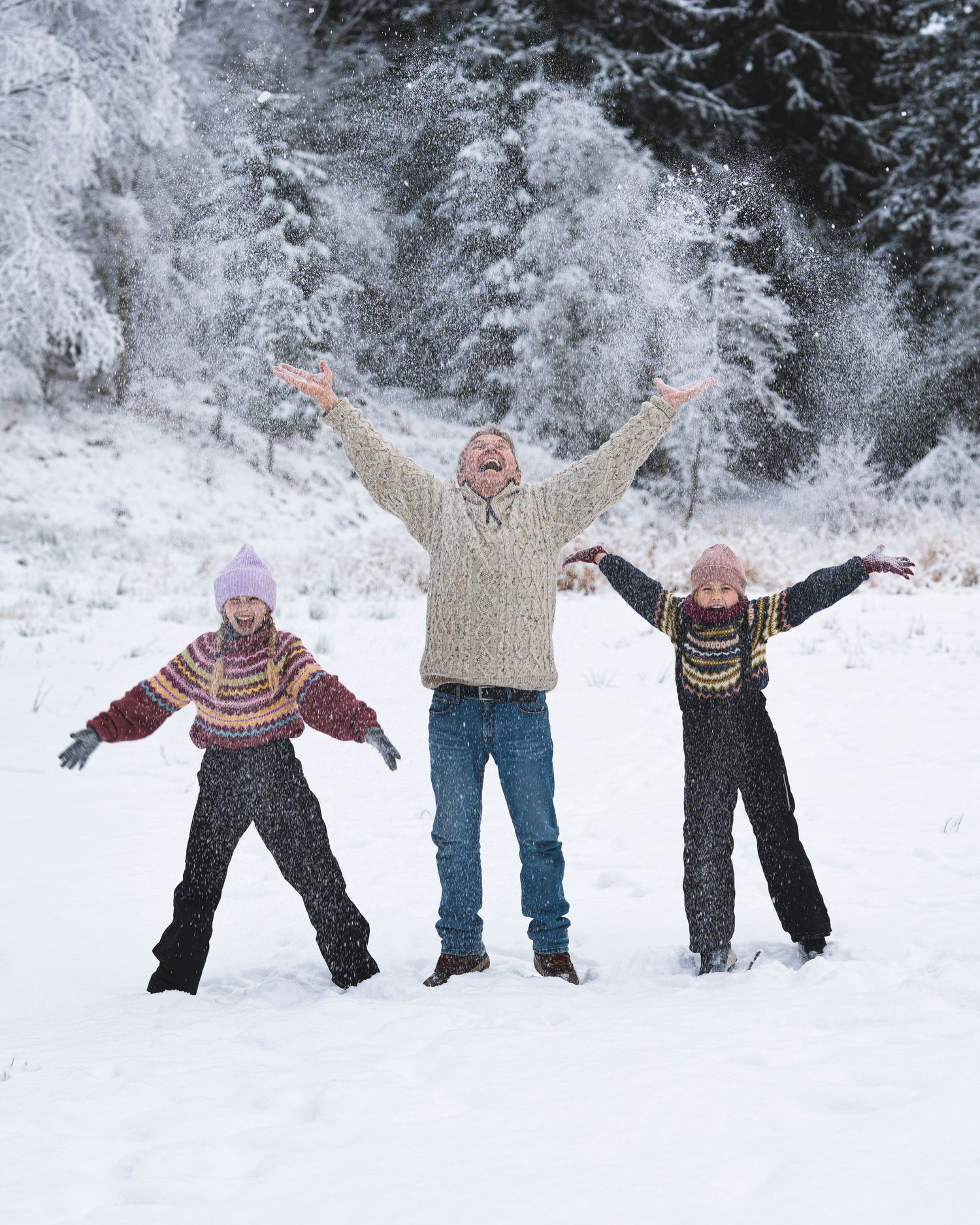 Three people play together in a snowy winter wonderland with arms outstretched.