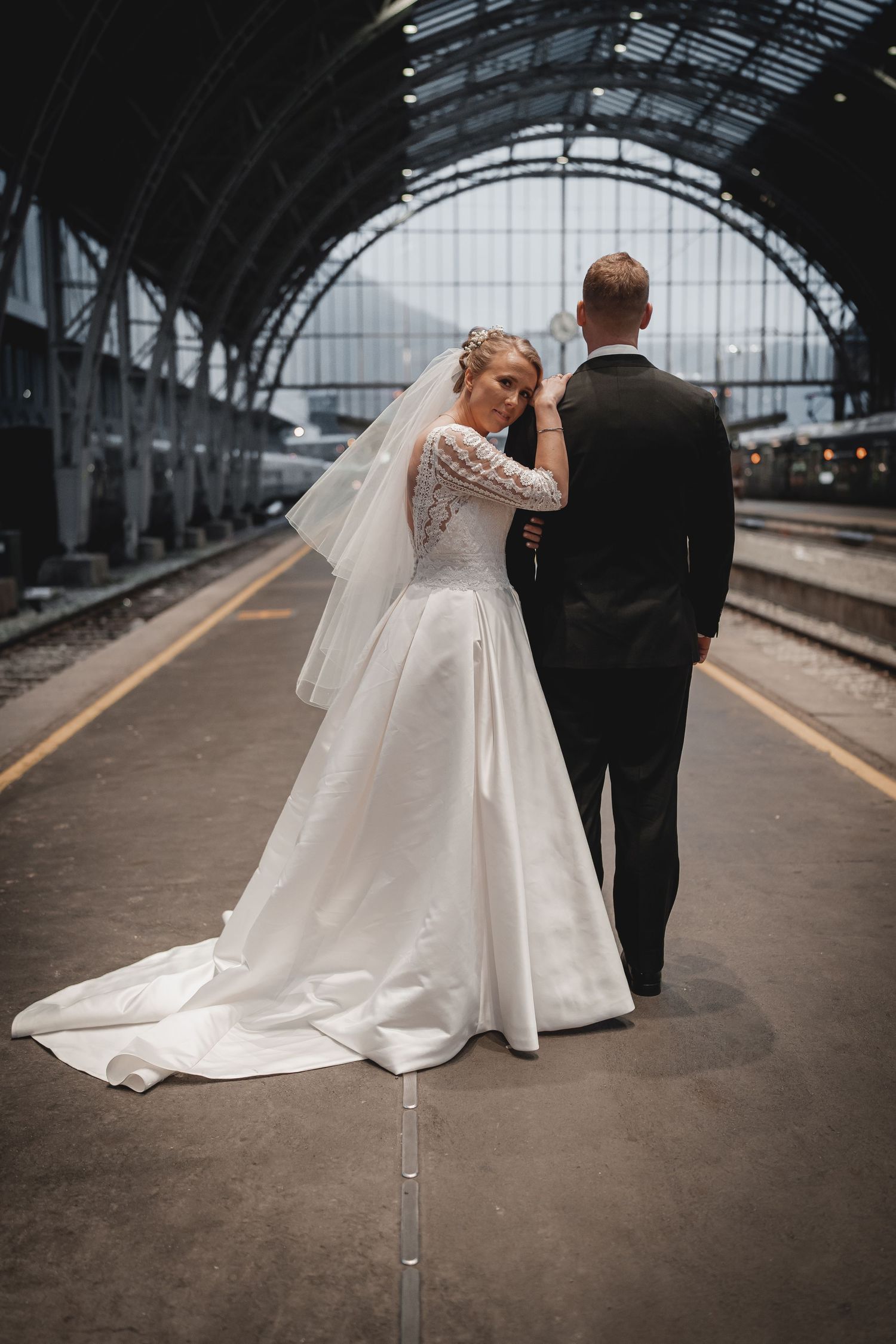 Bride in flowing white dress walking with partner in train station.