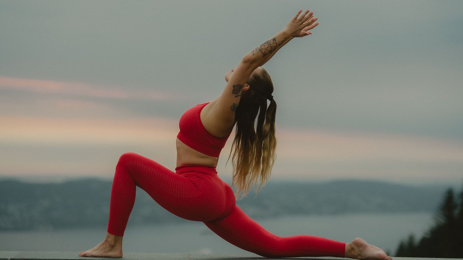 Kvinne i rødt treningstøy utfører yoga mot en vakker solnedgang ved kysten på Fløyen i Bergen