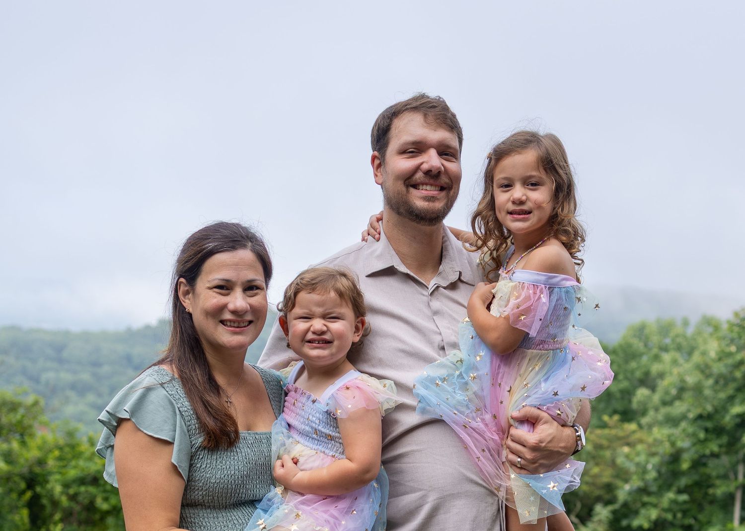 A happy family of four poses outdoors with lush green mountains in the background on a cloudy day.