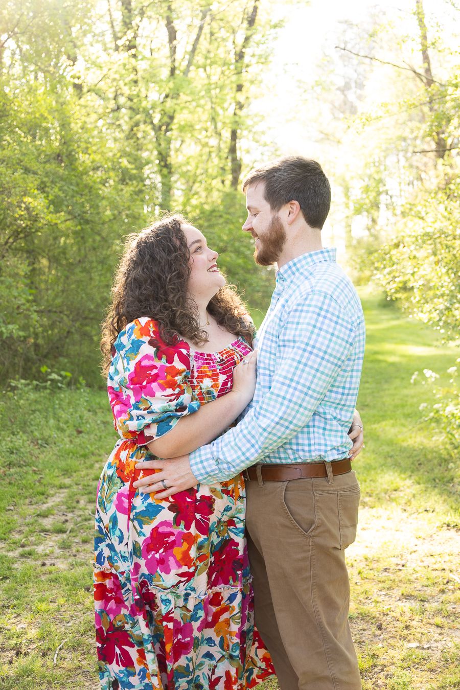 A couple embraces and smiles at each other outdoors, woman in floral dress, man in plaid shirt, surrounded by green trees.