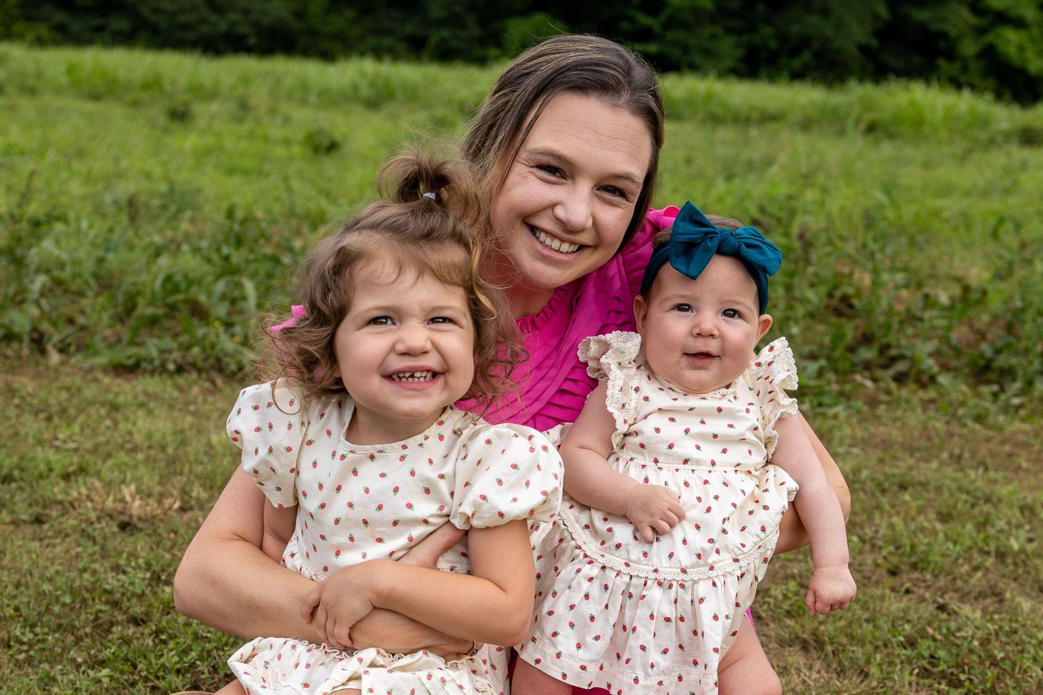 A smiling woman in pink holds two young girls in matching white floral dresses outdoors on green grass.