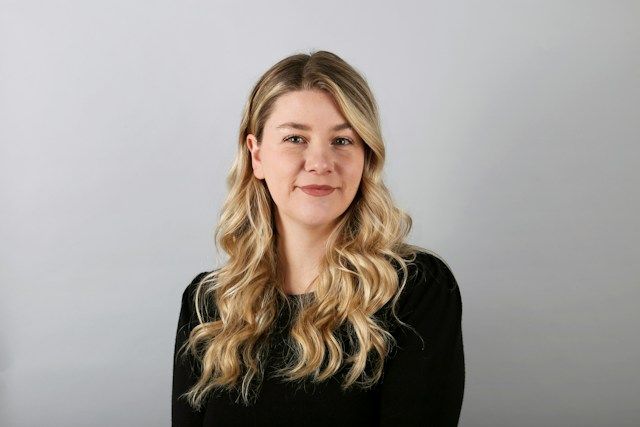 Professional corporate headshot of a blonde woman wearing a black top against a neutral background.