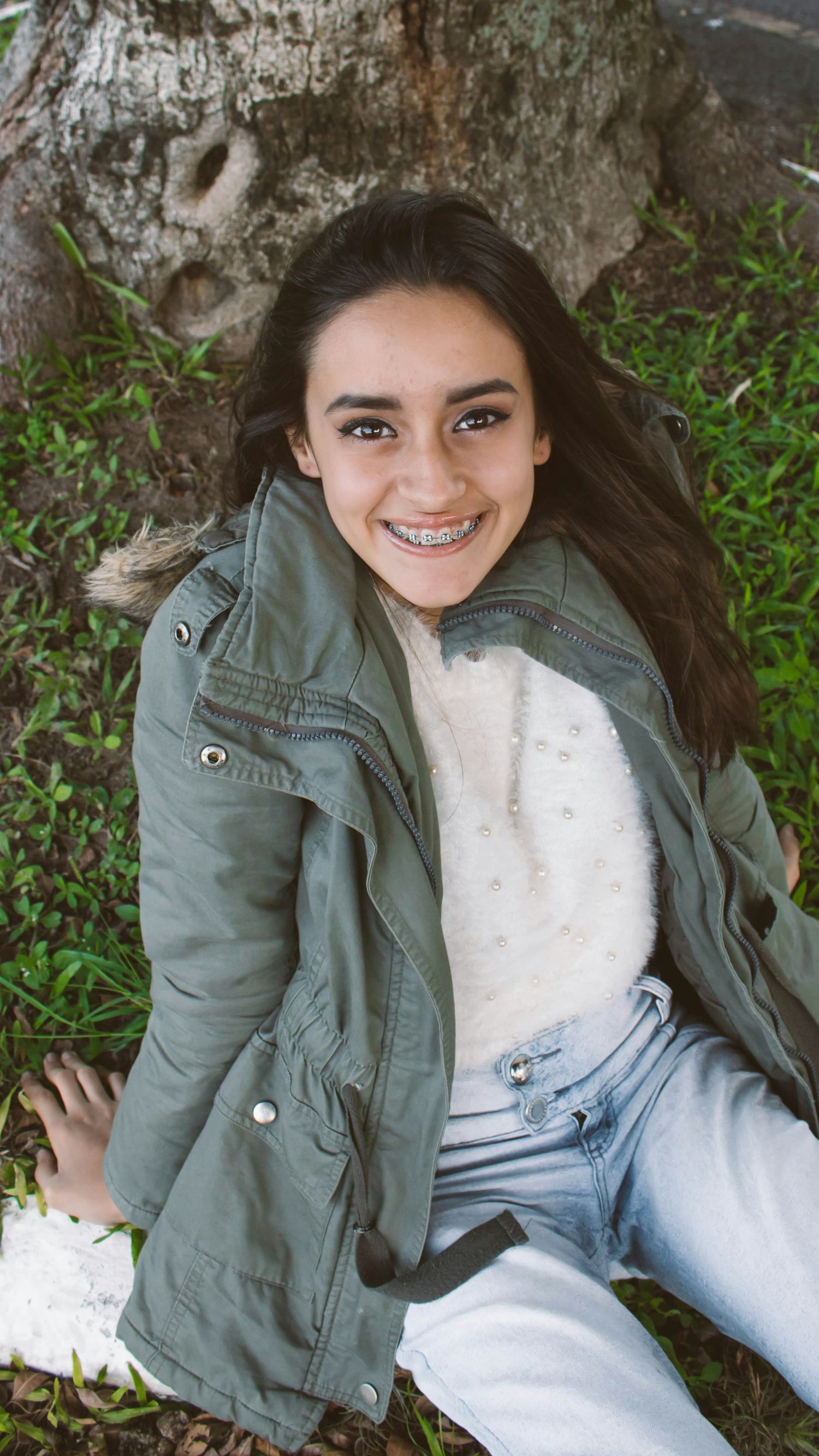 Smiling teenage girl with braces sitting against a tree, wearing a green jacket and jeans outdoors.