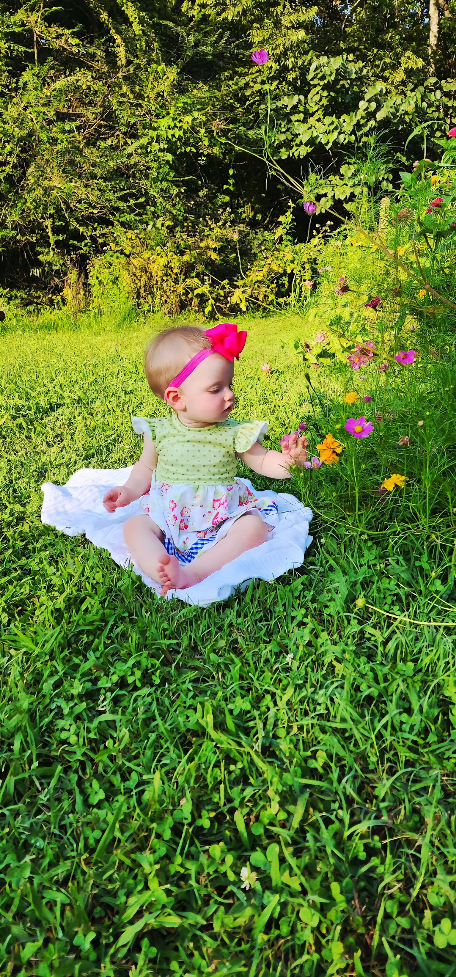Baby in floral dress lying on white blanket among wildflowers and grass.
