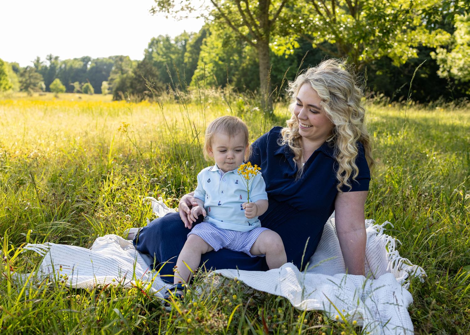 A smiling mother sits with her baby boy on a blanket in a sunny green meadow, the child holding a small flower.