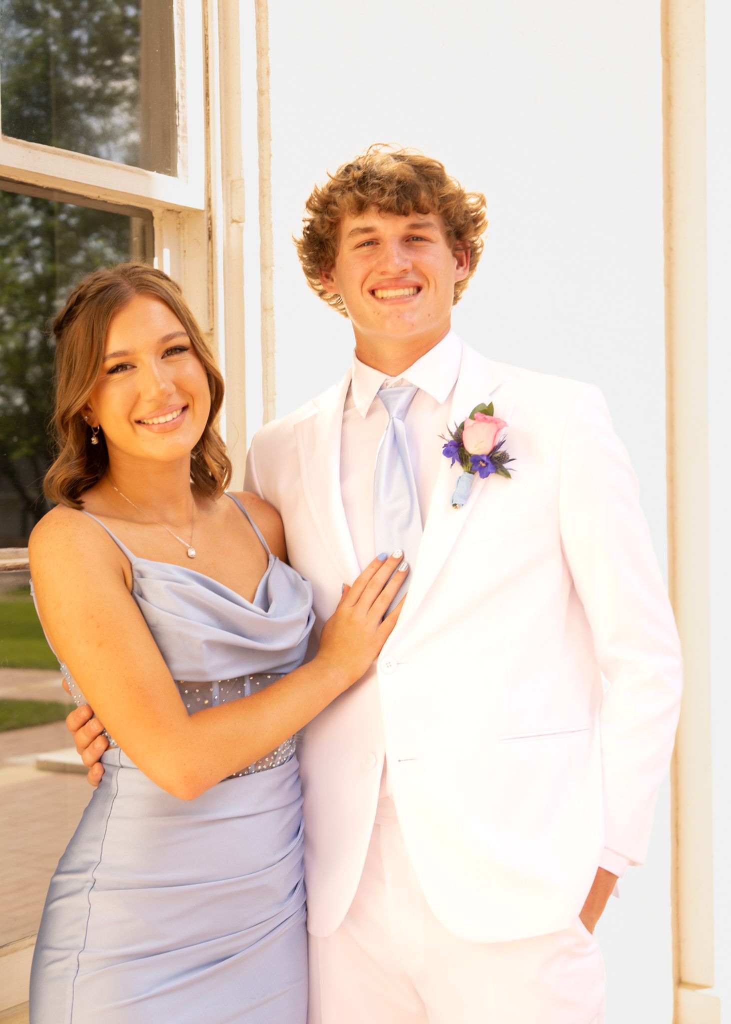 A smiling couple dressed for prom, girl in blue gown and boy in white suit with boutonniere, posing outdoors.