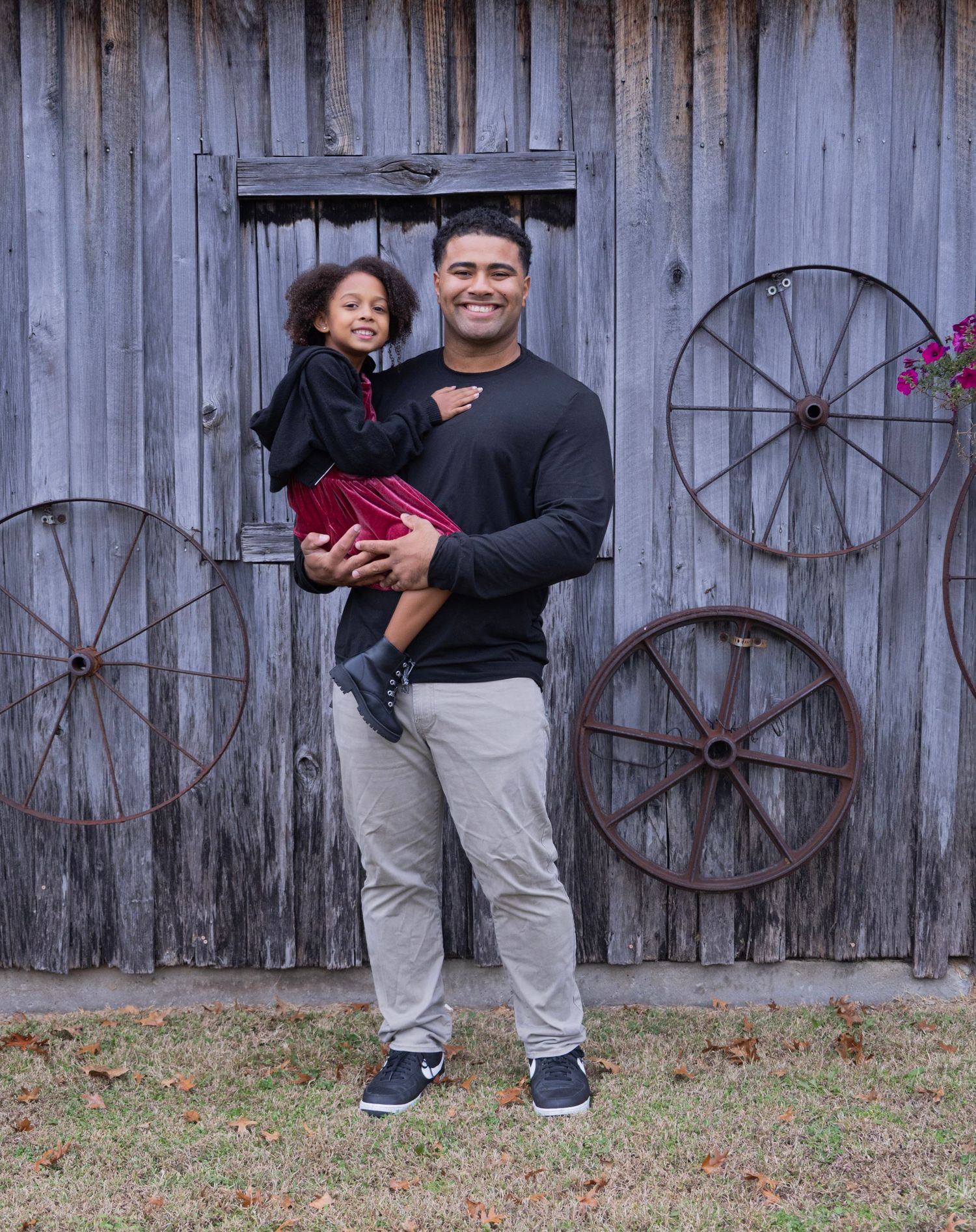 People stand in front of a rustic wooden barn wall decorated with vintage wagon wheels and pink flowers.