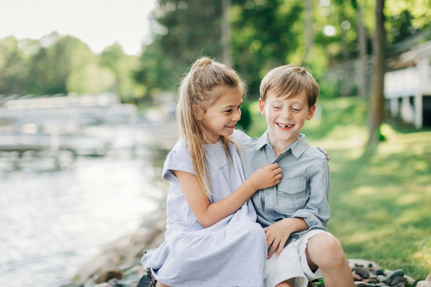 Two children sit together by a lakeside, a girl in a white dress and a boy in a gray shirt, smiling and laughing outdoors.