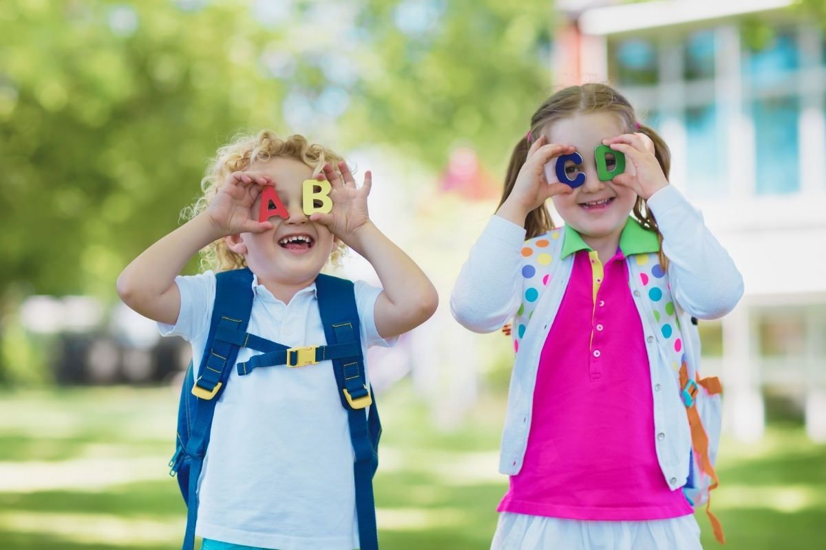 Two young children holding colorful alphabet letters over their eyes, wearing backpacks outdoors on a sunny day.