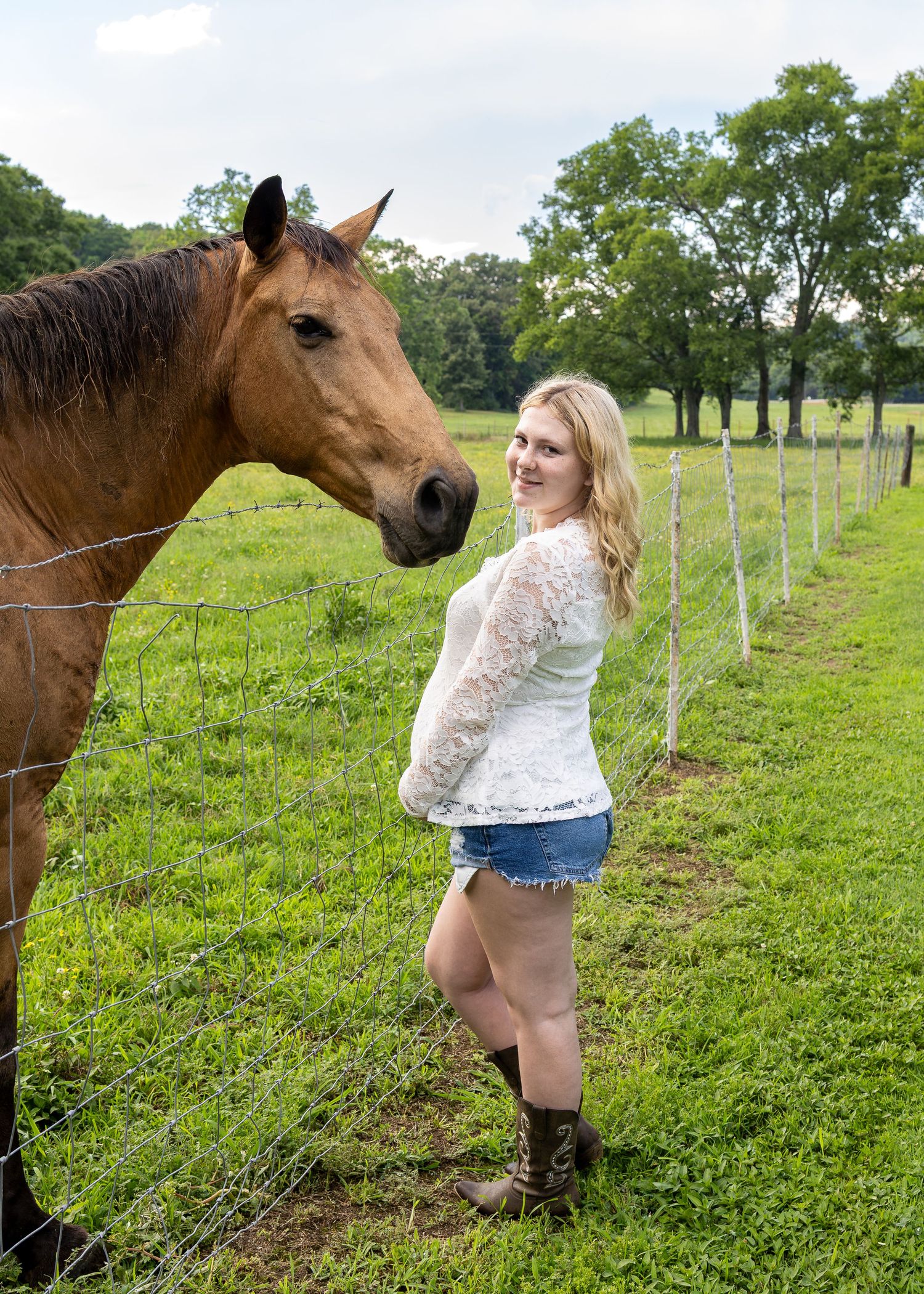 Blonde woman in white lace top and denim shorts stands by fence with brown horse on green farm.