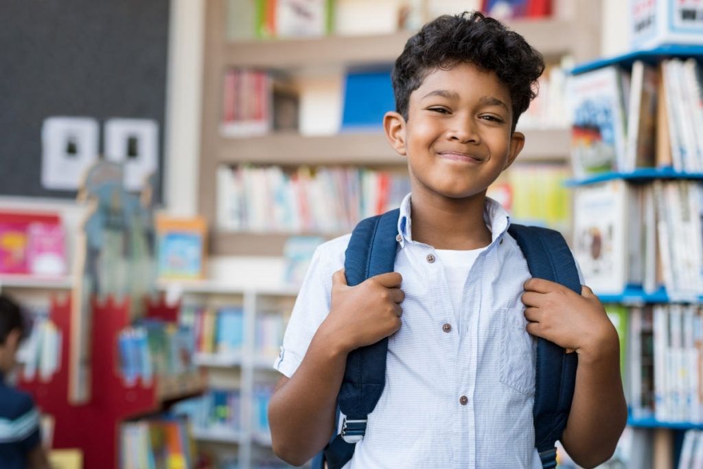 Smiling young boy with backpack standing in a school library, ready for learning.