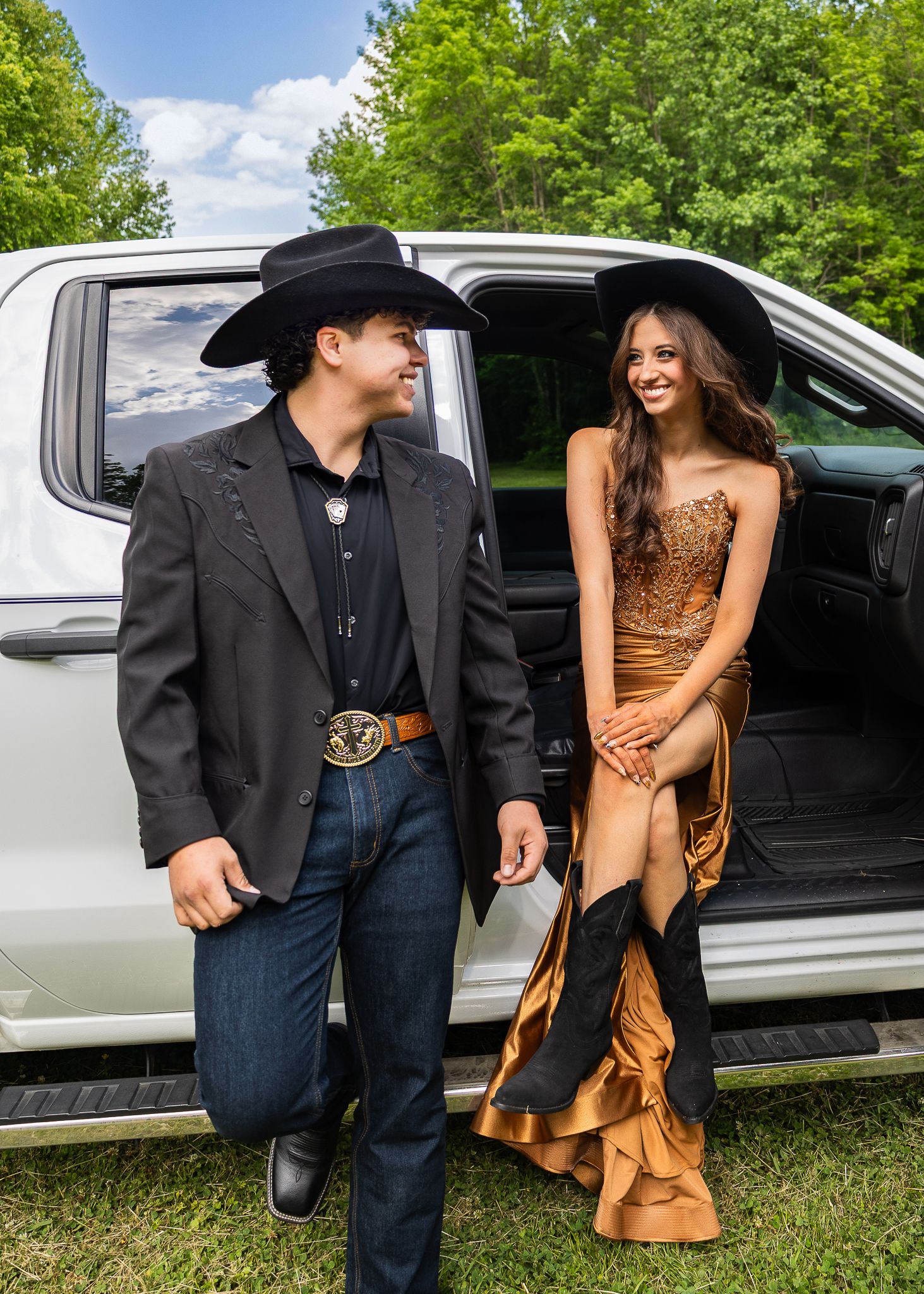 Couple in Western attire, man in black cowboy hat and blazer, woman in gold gown, posing by a white truck outdoors.