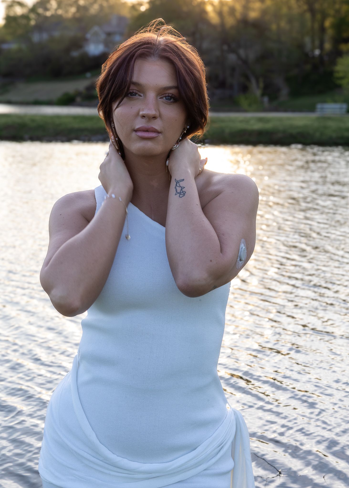 Young woman in white dress posing near a reflective lake at sunset, with tattoos and auburn hair.