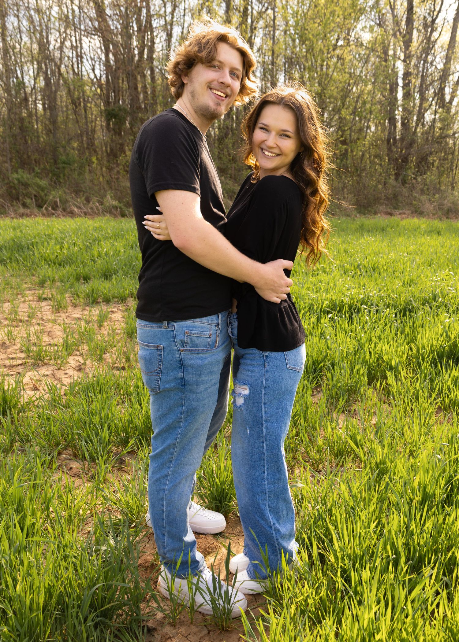 A smiling couple in matching black tops and jeans embracing outdoors in a lush green field with trees in the background.