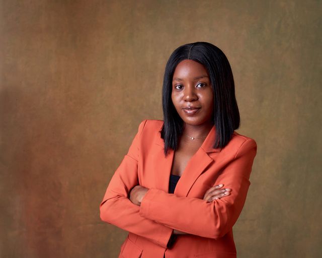Professional woman in orange blazer posing confidently with arms crossed against a brown studio background.