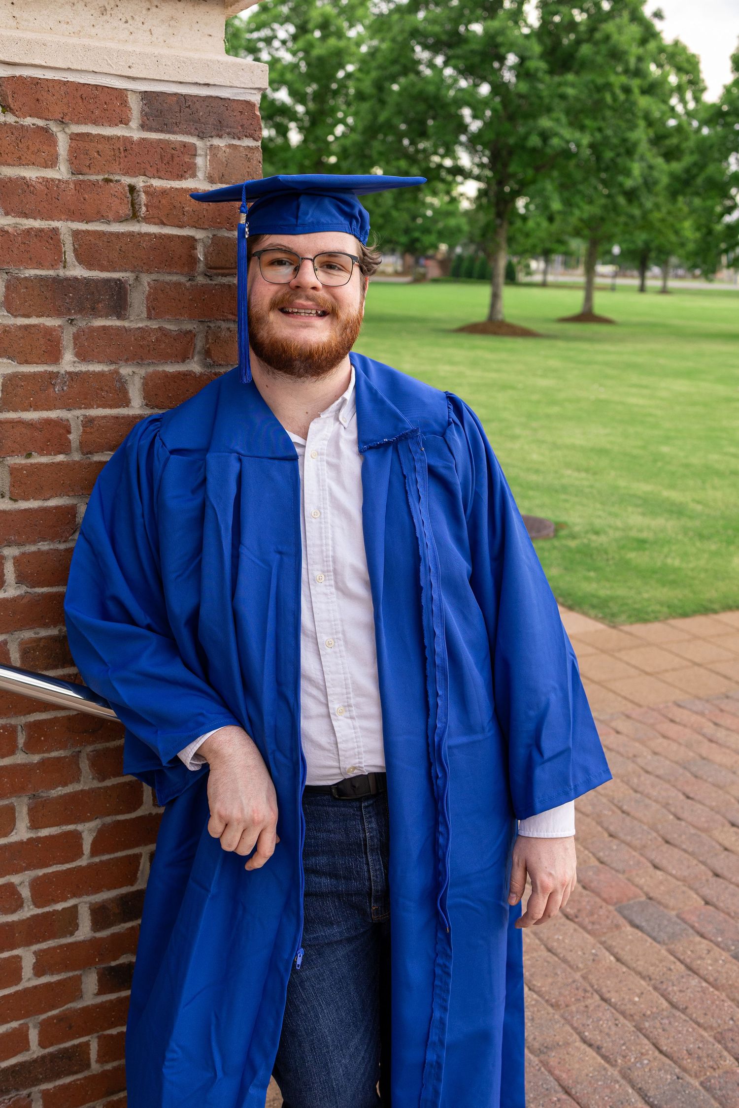 Young man smiling in blue graduation cap and gown, leaning against brick wall on green campus grounds.