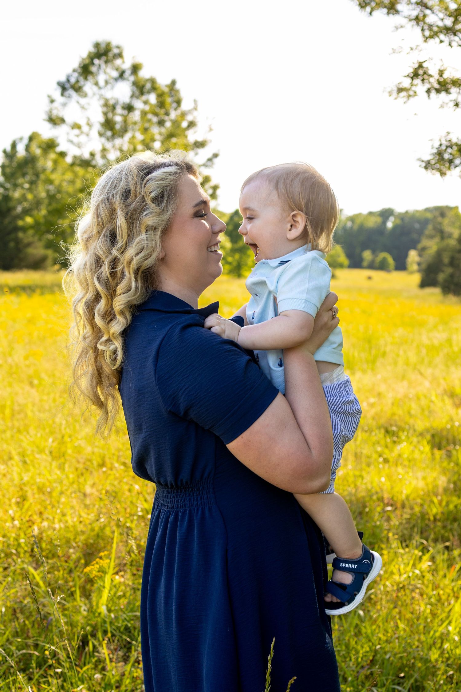 A smiling blonde woman in a navy dress holds a happy baby boy in a sunny yellow wildflower field.