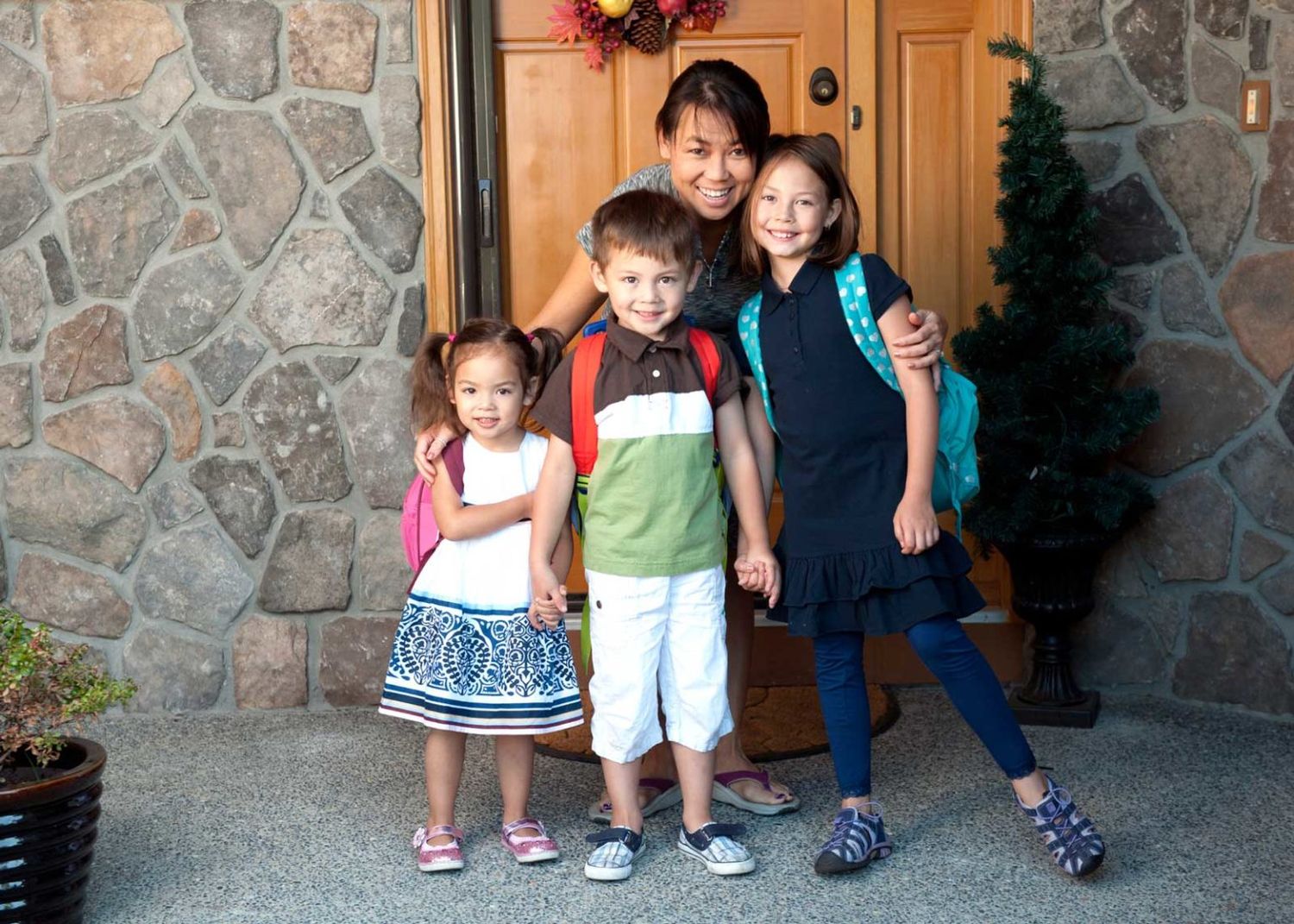 A woman poses with three children wearing backpacks in front of a stone house entrance on the first day of school.